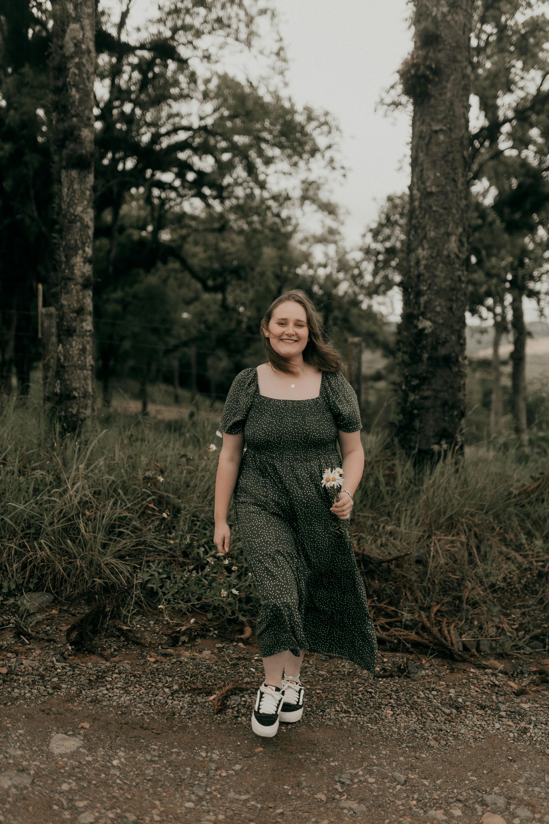 Woman in green dress walks toward camera on a dirt path, holding flowers. Trees in background.