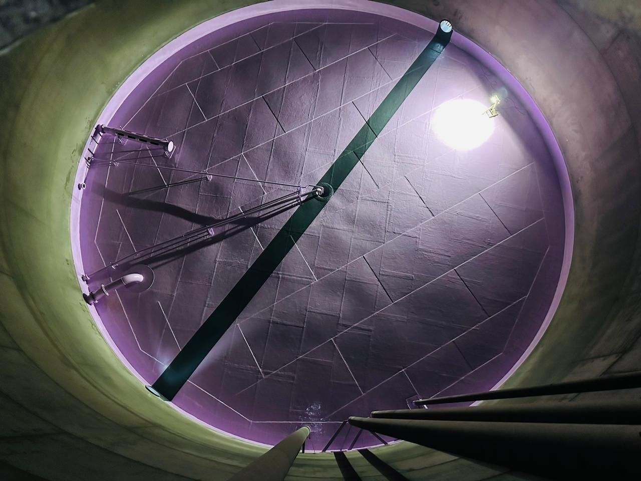 Interior de um túnel circular iluminado de roxo, com um feixe diagonal verde e uma luz brilhante perto do topo.