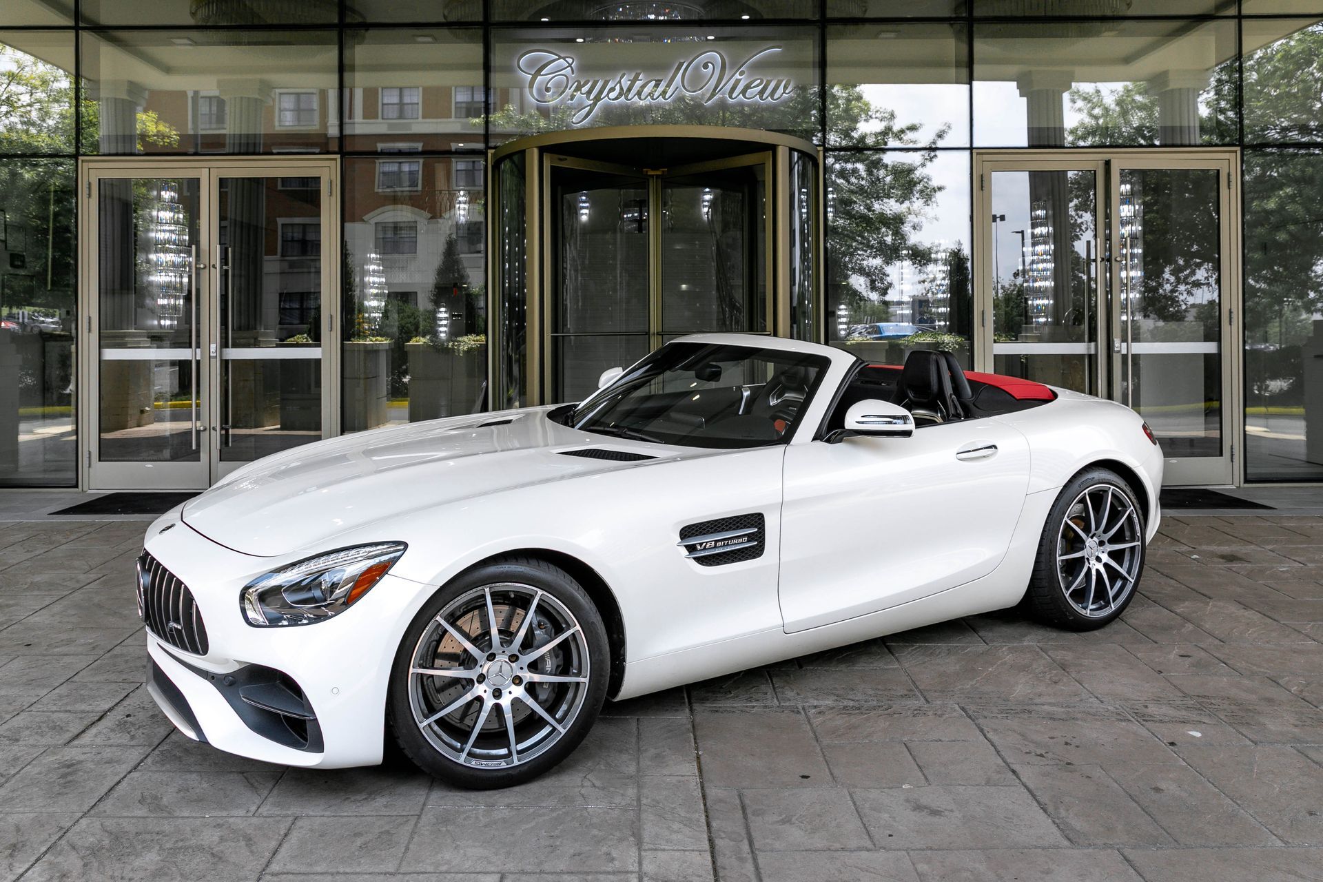 White convertible sports car parked in front of a building with a revolving door; the car's top is down, red interior.
