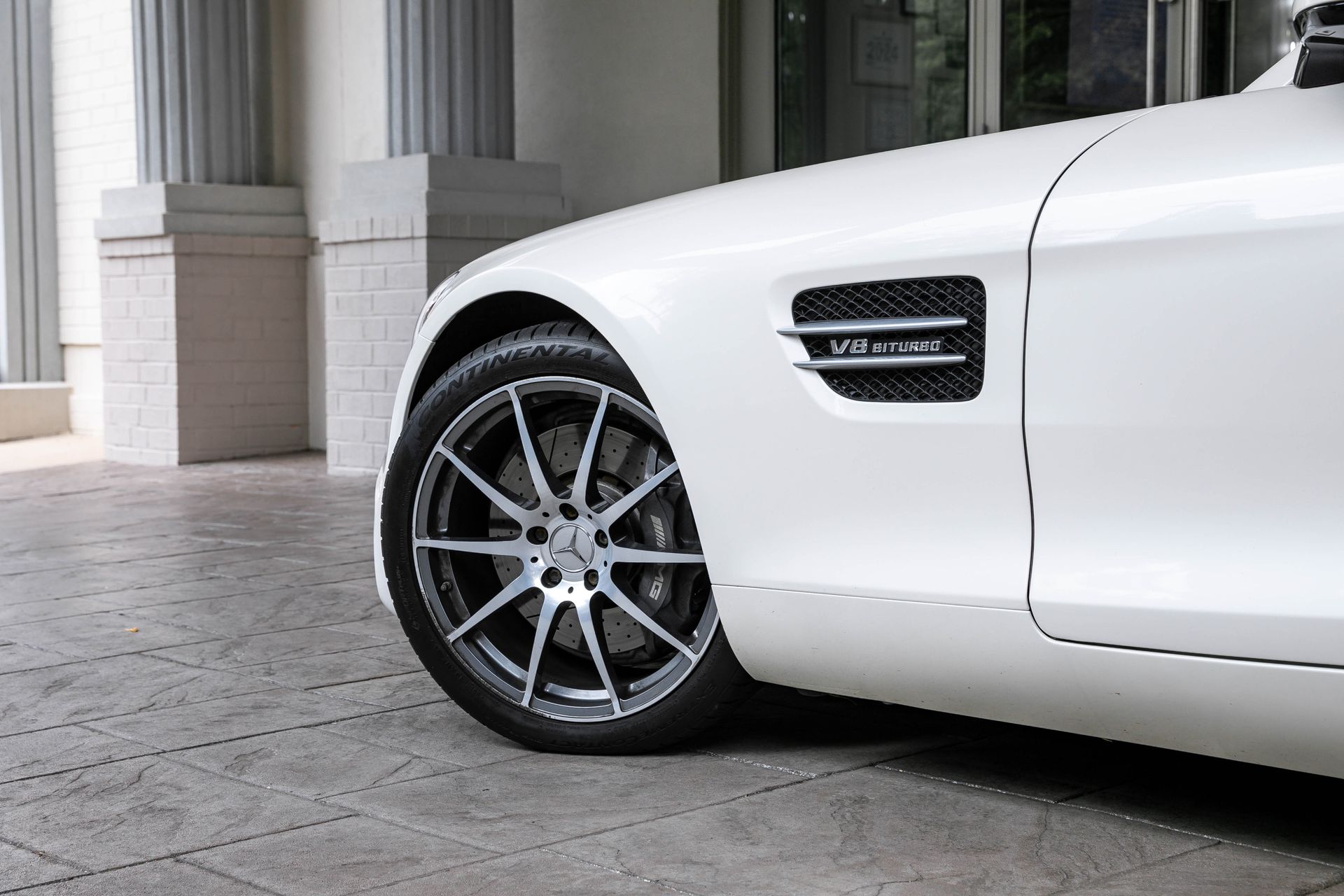 White Mercedes-AMG GT sports car, front quarter view, with a black and silver wheel, parked near a building.