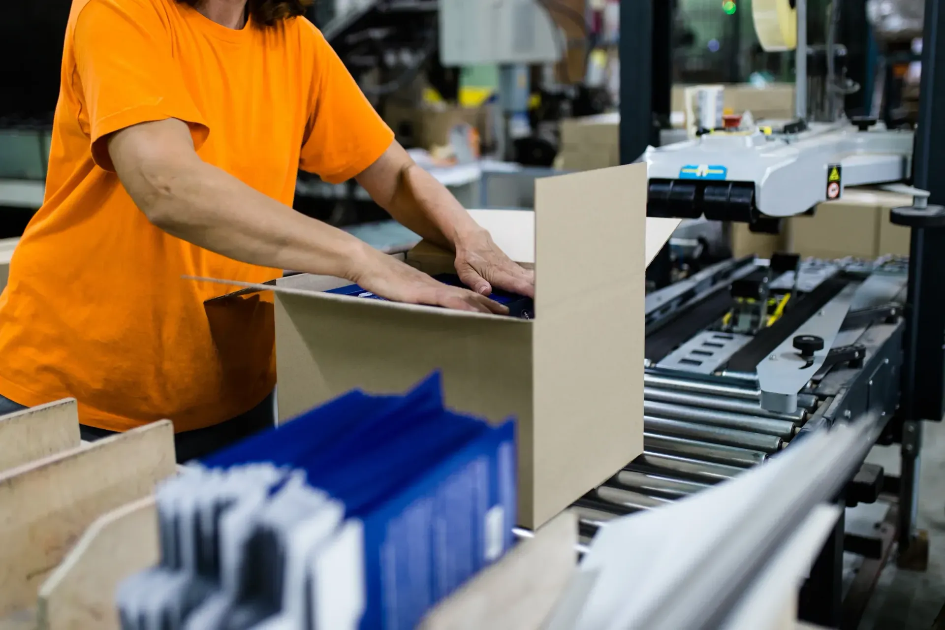Woman in orange shirt packing a cardboard box on a conveyor belt in a factory.