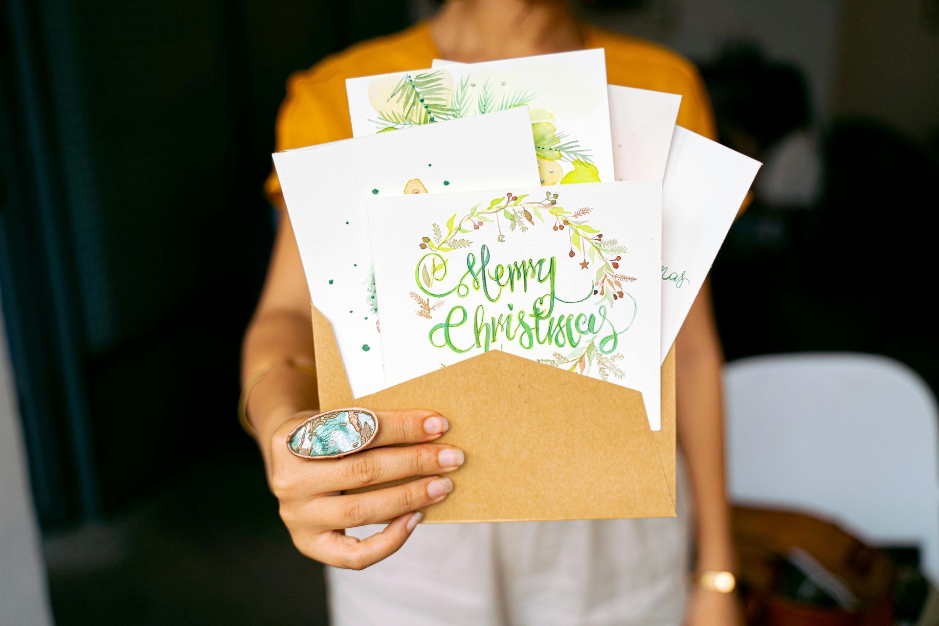Person holding an open brown envelope with several watercolor Christmas cards; the central card says 
