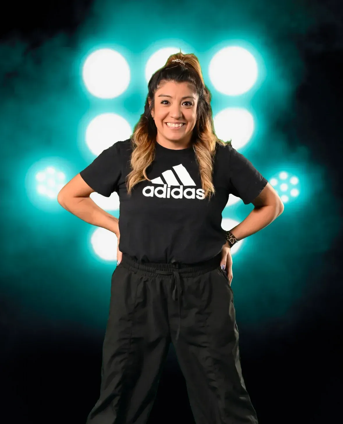 Woman in black Adidas shirt and pants, hands on hips, smiles in front of stage lights.