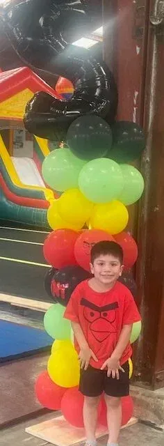 Boy in red shirt smiles, poses by a column of colorful balloons. Indoors, with bouncy castle in background.