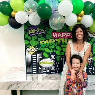 Woman and child smiling, posed in front of a superhero-themed birthday backdrop with balloons.