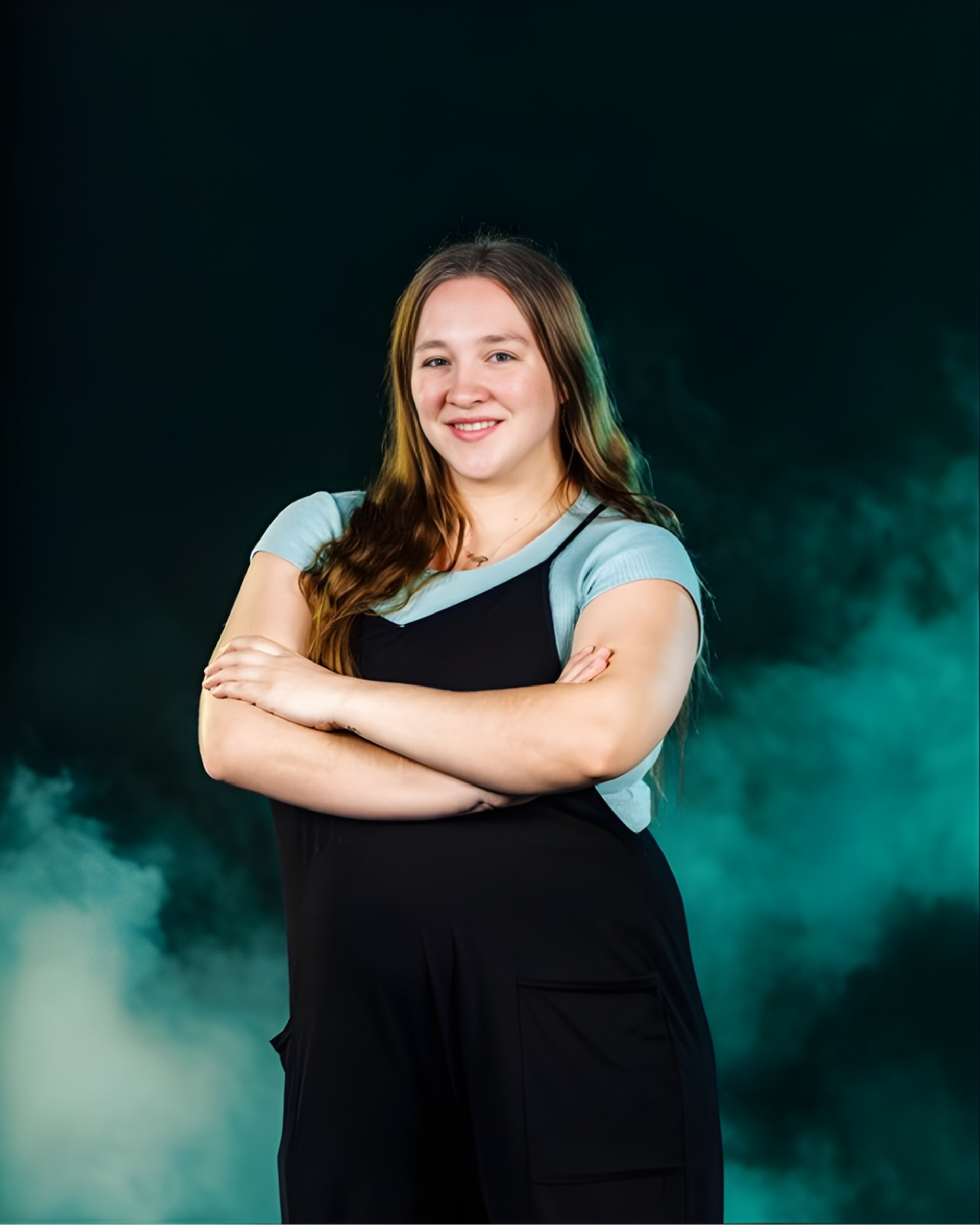 A person with long hair smiles with arms crossed, wearing a black jumpsuit over a light blue shirt against a teal fog.