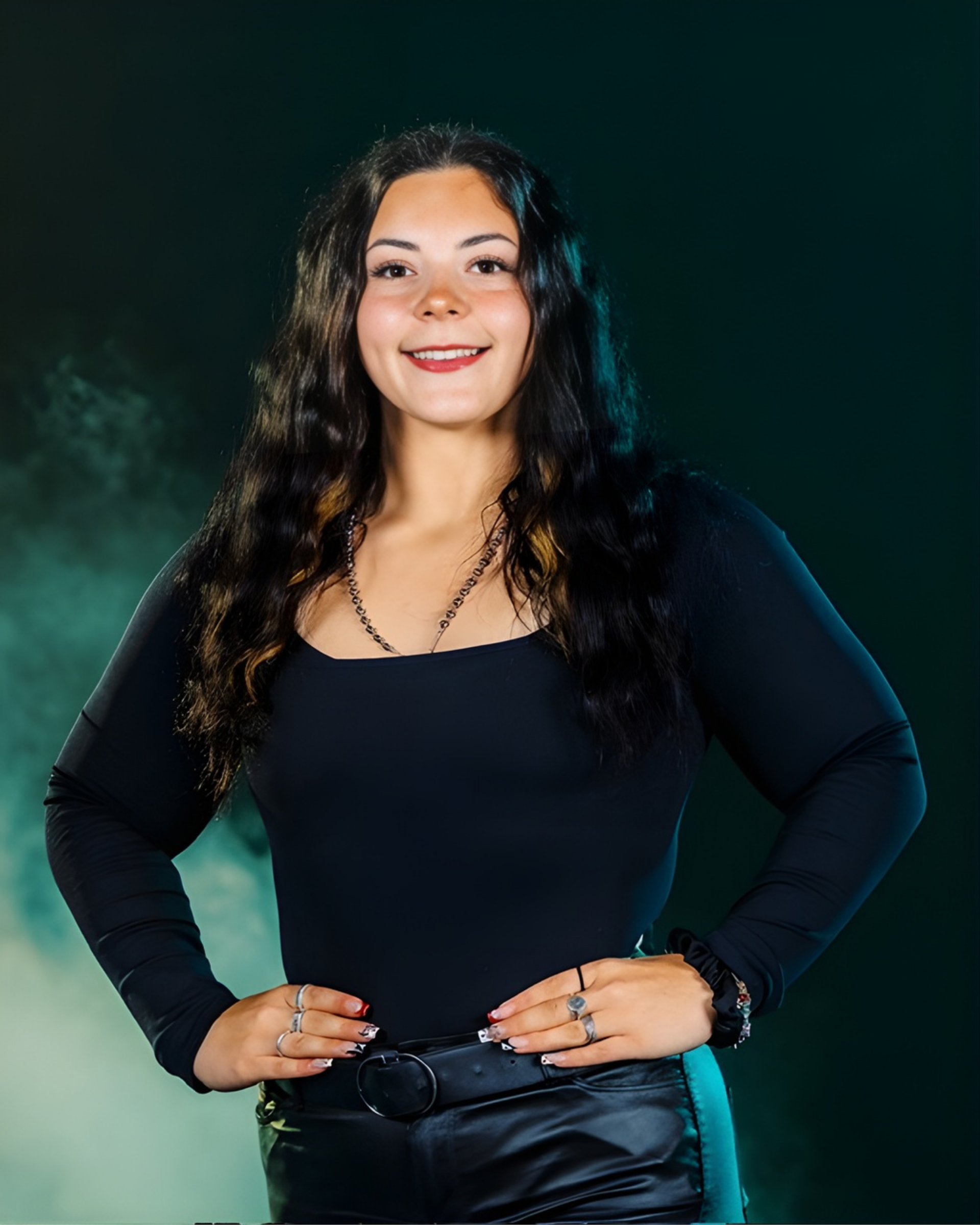 A person with long dark hair, wearing a black long-sleeve top and belt, poses with hands on hips against a smoky background.
