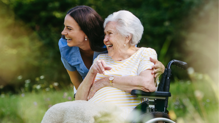 Woman in blue shirt embraces elderly person in wheelchair outdoors; both smiling.