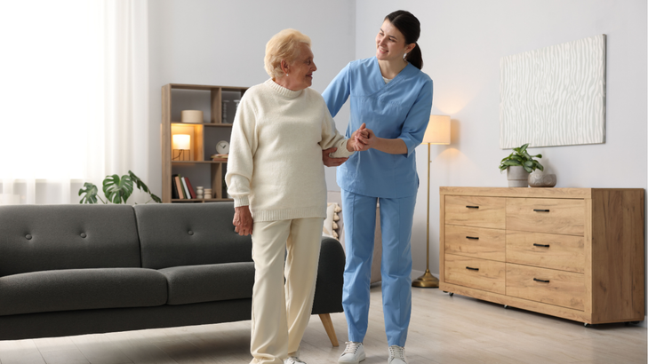 Caregiver assists elderly person walking in a living room.