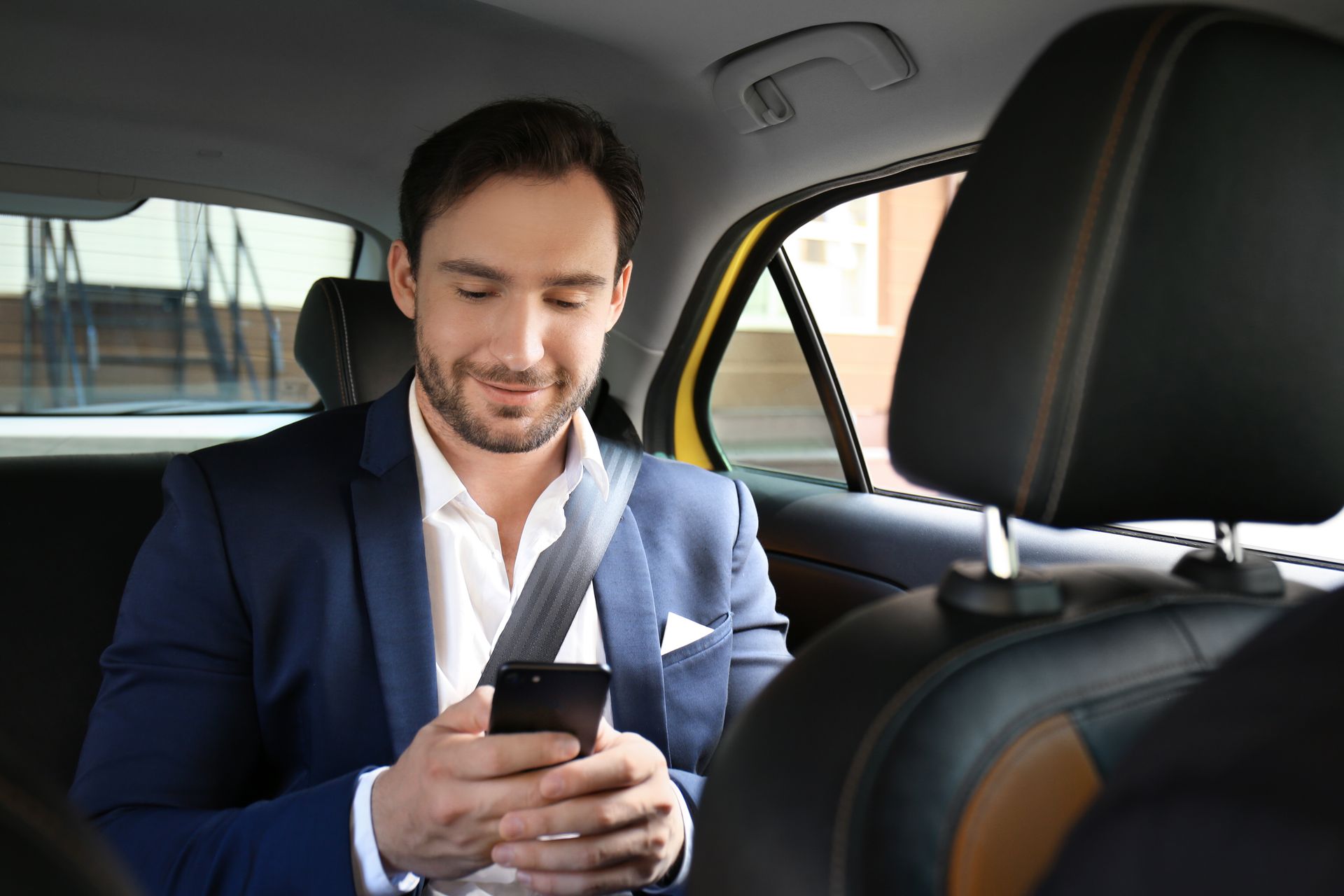 A man in a suit is sitting in the back seat of a car looking at his cell phone.