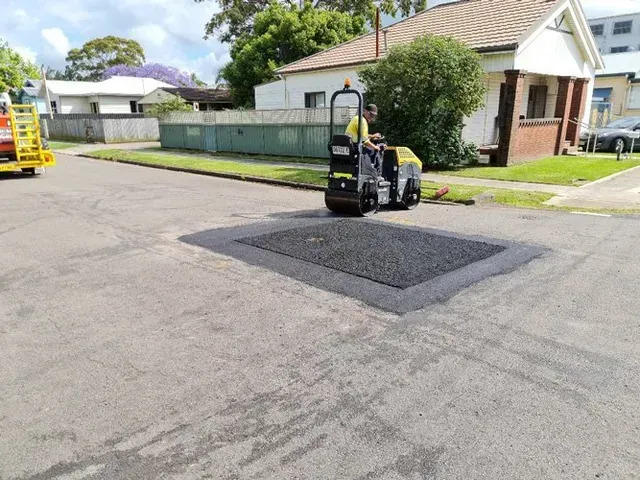 A construction worker operates a yellow asphalt roller to smooth a freshly paved rectangular patch on a suburban road.