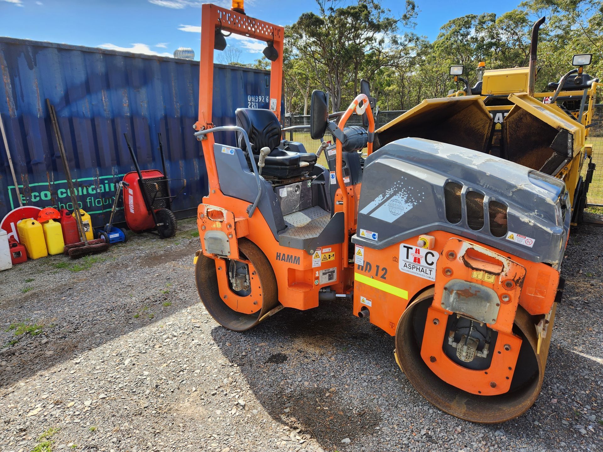 Orange And Gray Road Roller On Gravel With A Dark Blue Fence — TAC Asphalt Services in Warners Bay, NSW