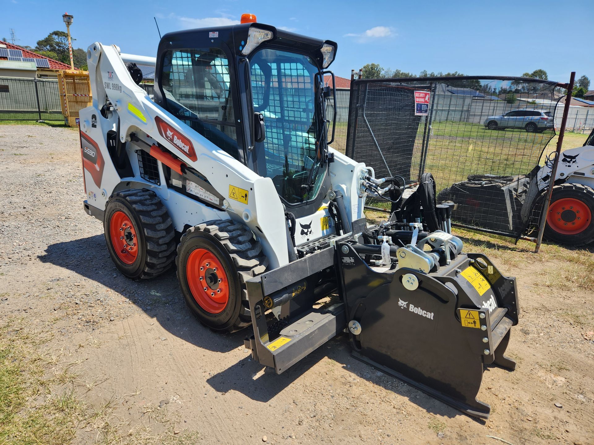 A white Bobcat skid-steer loader with orange wheels and a black industrial attachment parked on a gravel lot.