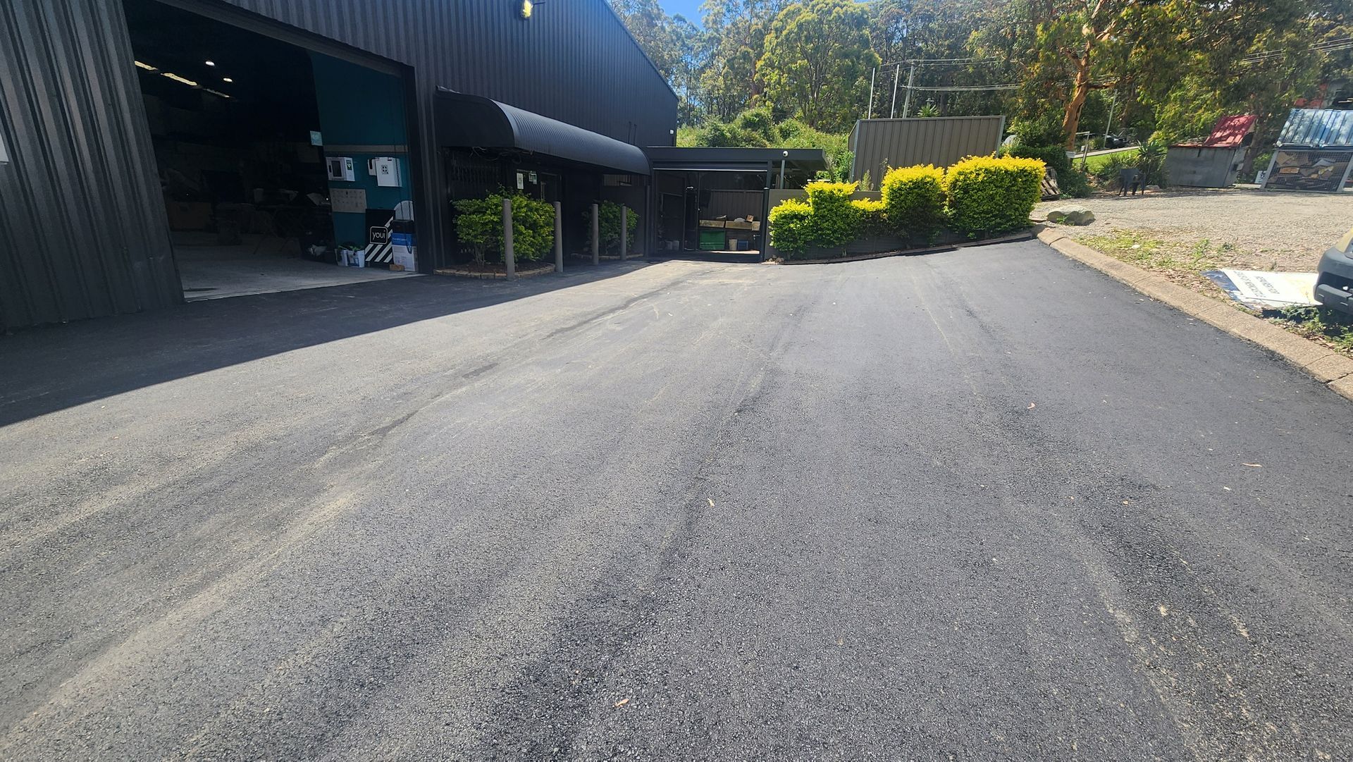 Asphalt Driveway Leading To A Dark Building With A Black Awning And Green Bushes — TAC Asphalt Services in Warners Bay, NSW
