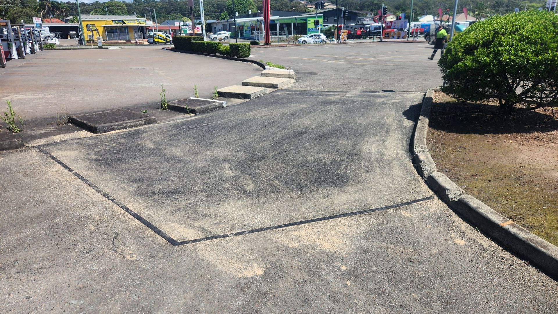 Asphalt Surface With a Damaged Patch, Curb, and Shrub in Front of a Building — TAC Asphalt Services in Warners Bay, NSW