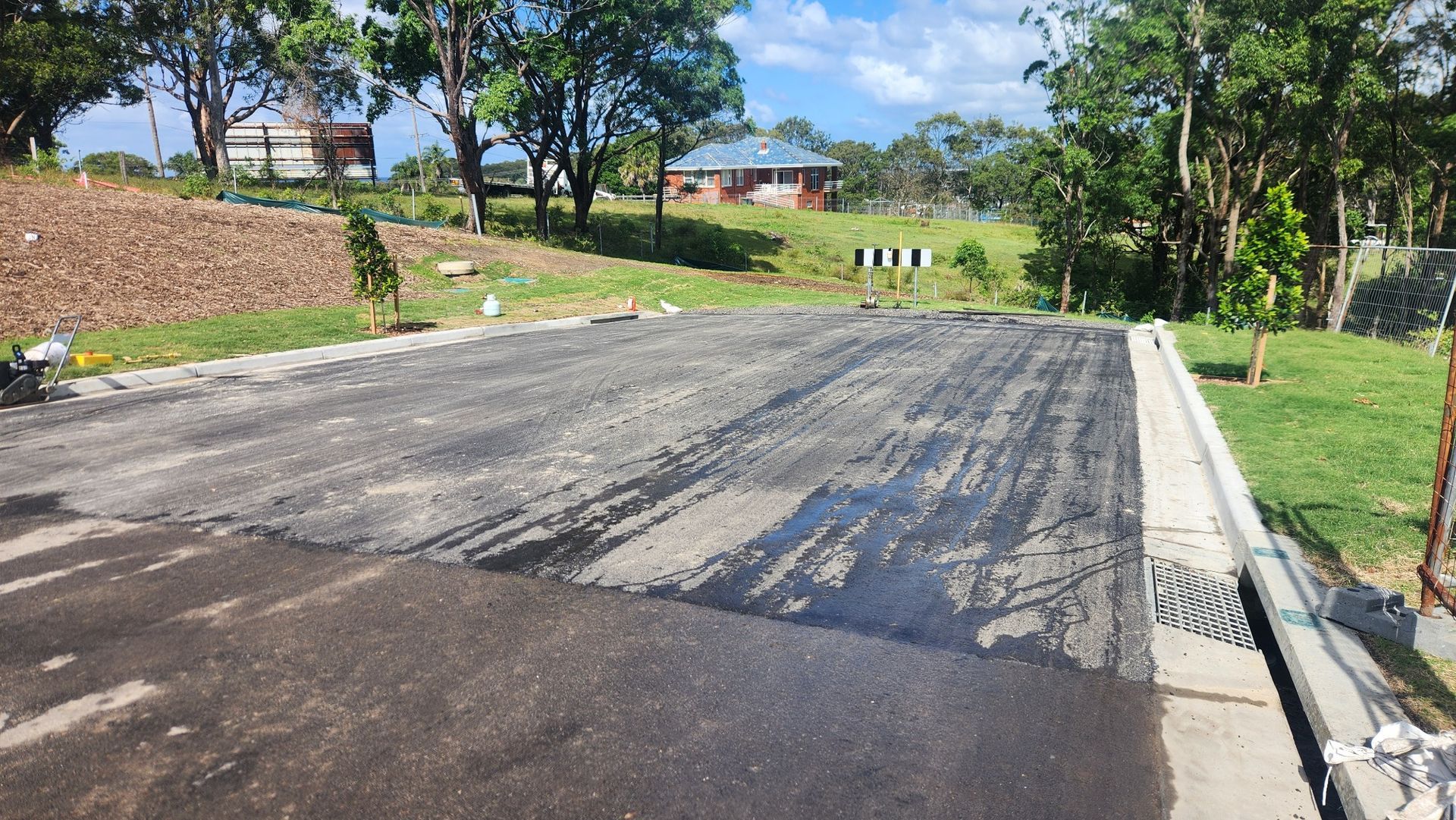 A newly paved asphalt road section with concrete curbs and a drainage grate, surrounded by green grass and trees.