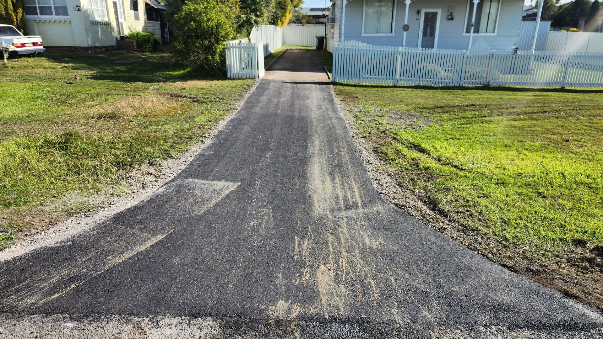 A newly laid dark asphalt driveway leading to a light-colored house, flanked by grass and a white fence.