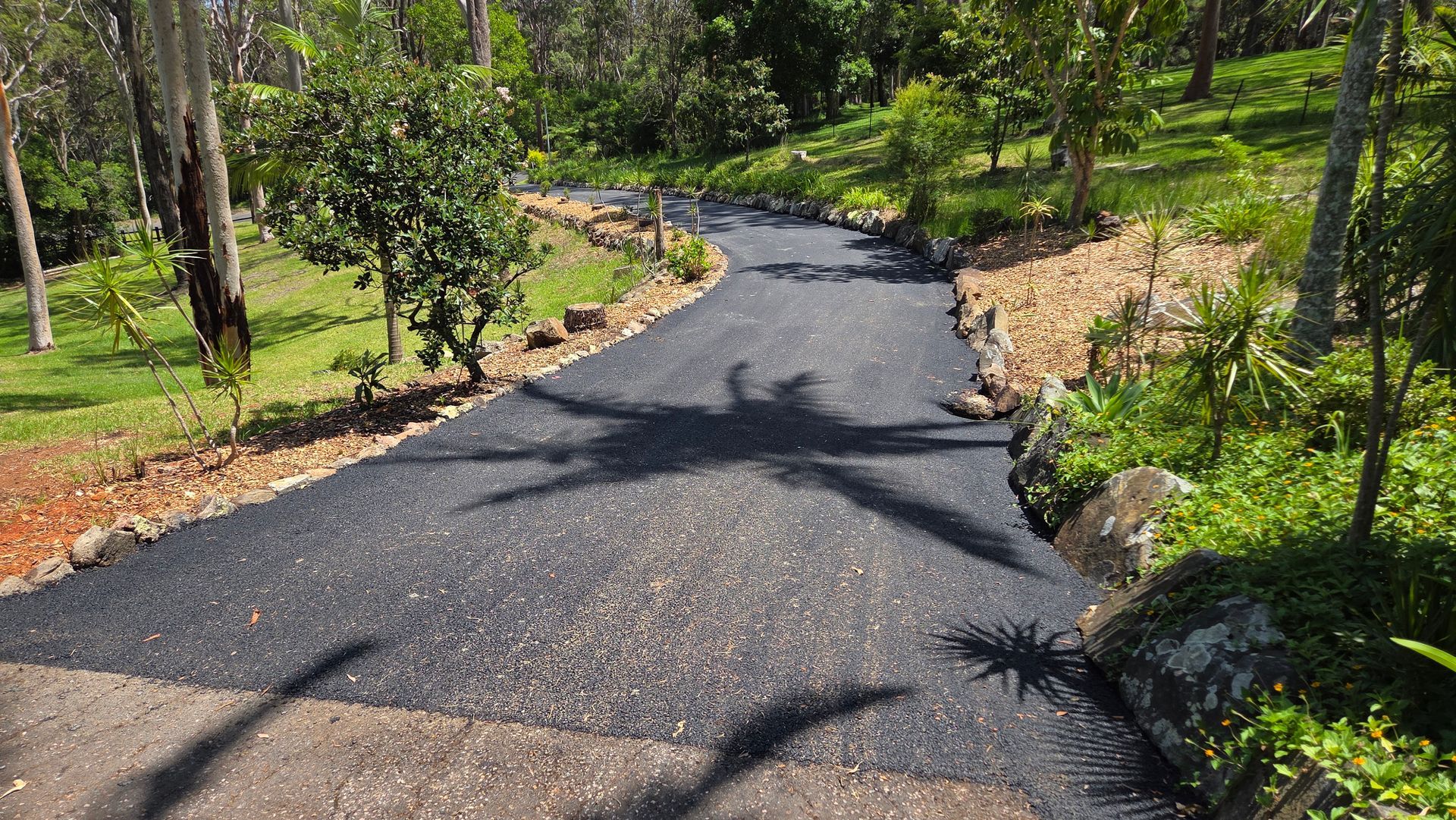 A dark asphalt driveway curves through a lush, green wooded landscape with dappled sunlight.