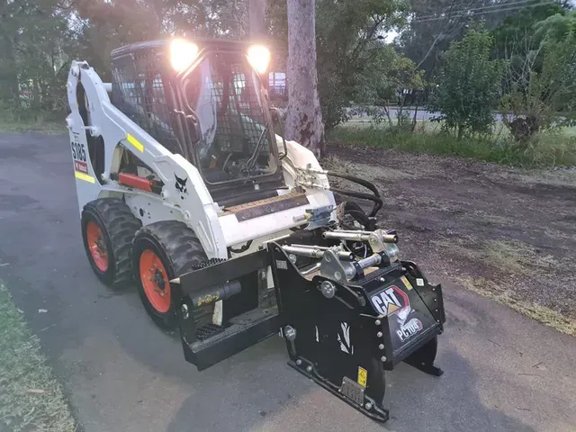 A white Bobcat skid-steer loader with a black CAT brand stump grinder attachment parked on a paved driveway at dusk.