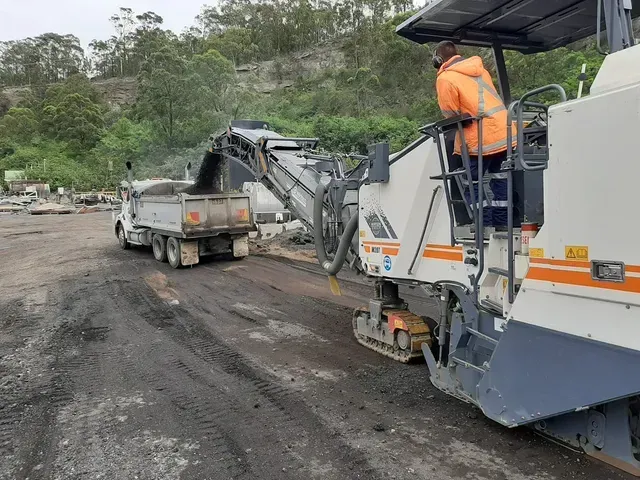 A worker in a high-visibility vest operates a road milling machine as it loads removed asphalt into a dump truck.