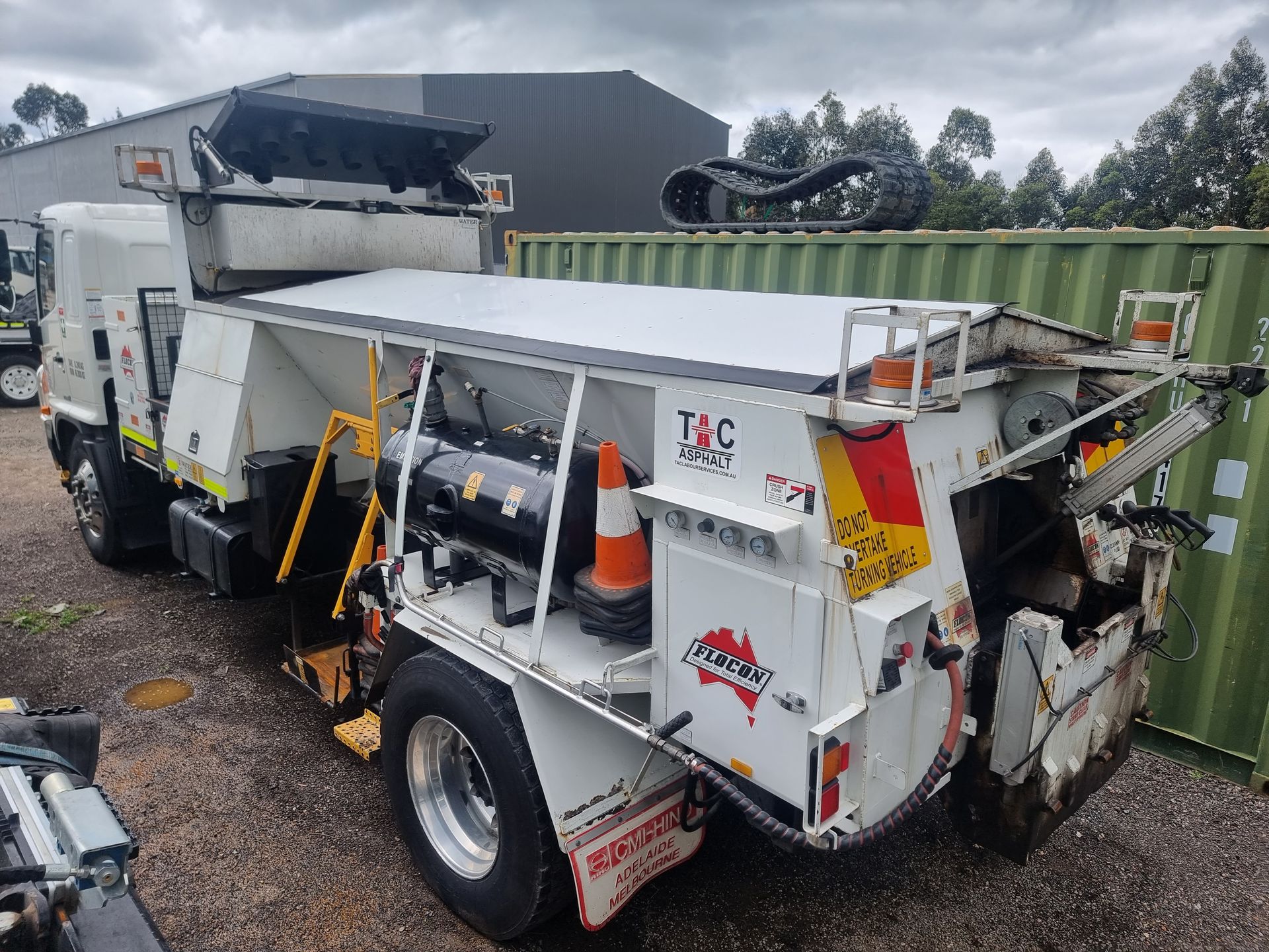 A white T&C road maintenance truck parked on a gravel lot next to a green shipping container.