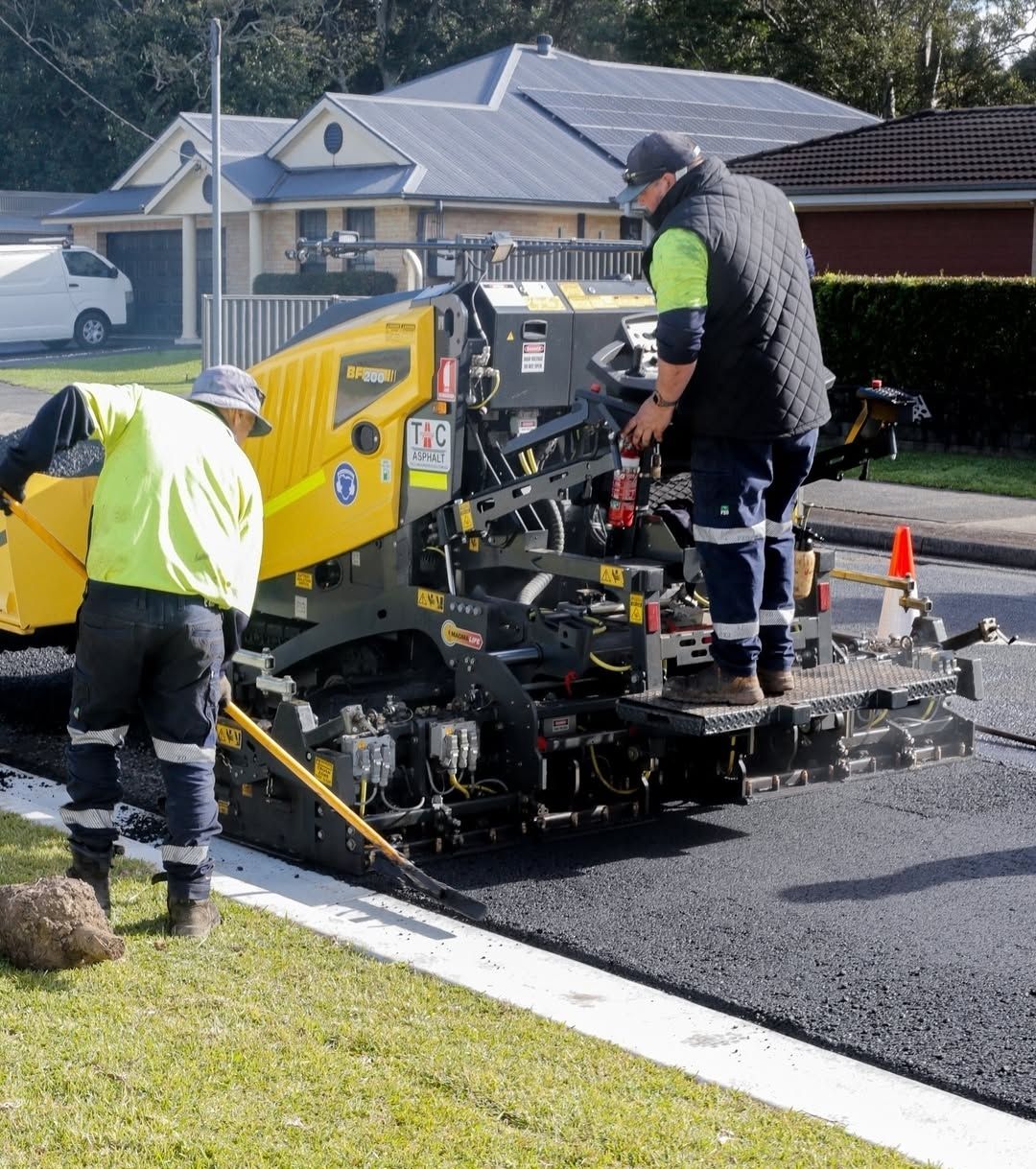 Two construction workers in high-visibility gear operate a yellow paving machine to lay asphalt on a residential street.