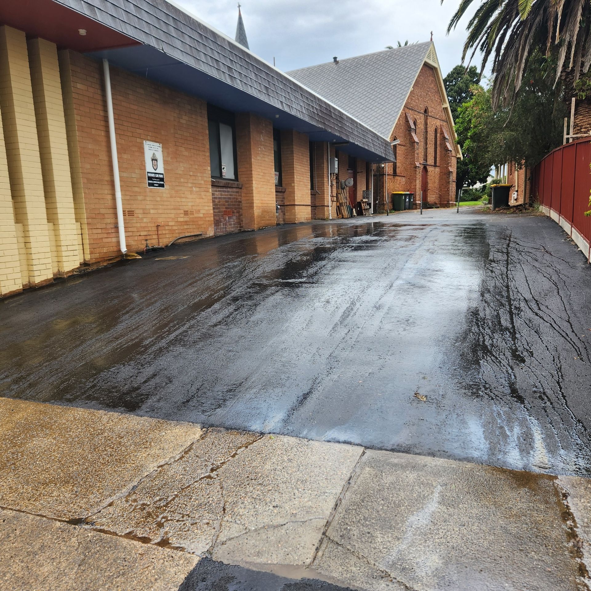 A wet, newly paved black asphalt driveway runs between a modern brick building and a historic stone church.