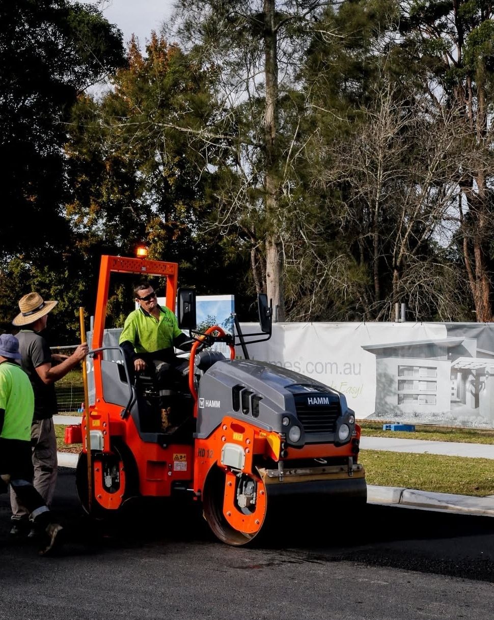 A worker operates an orange road roller to pave asphalt on a street near a construction site.