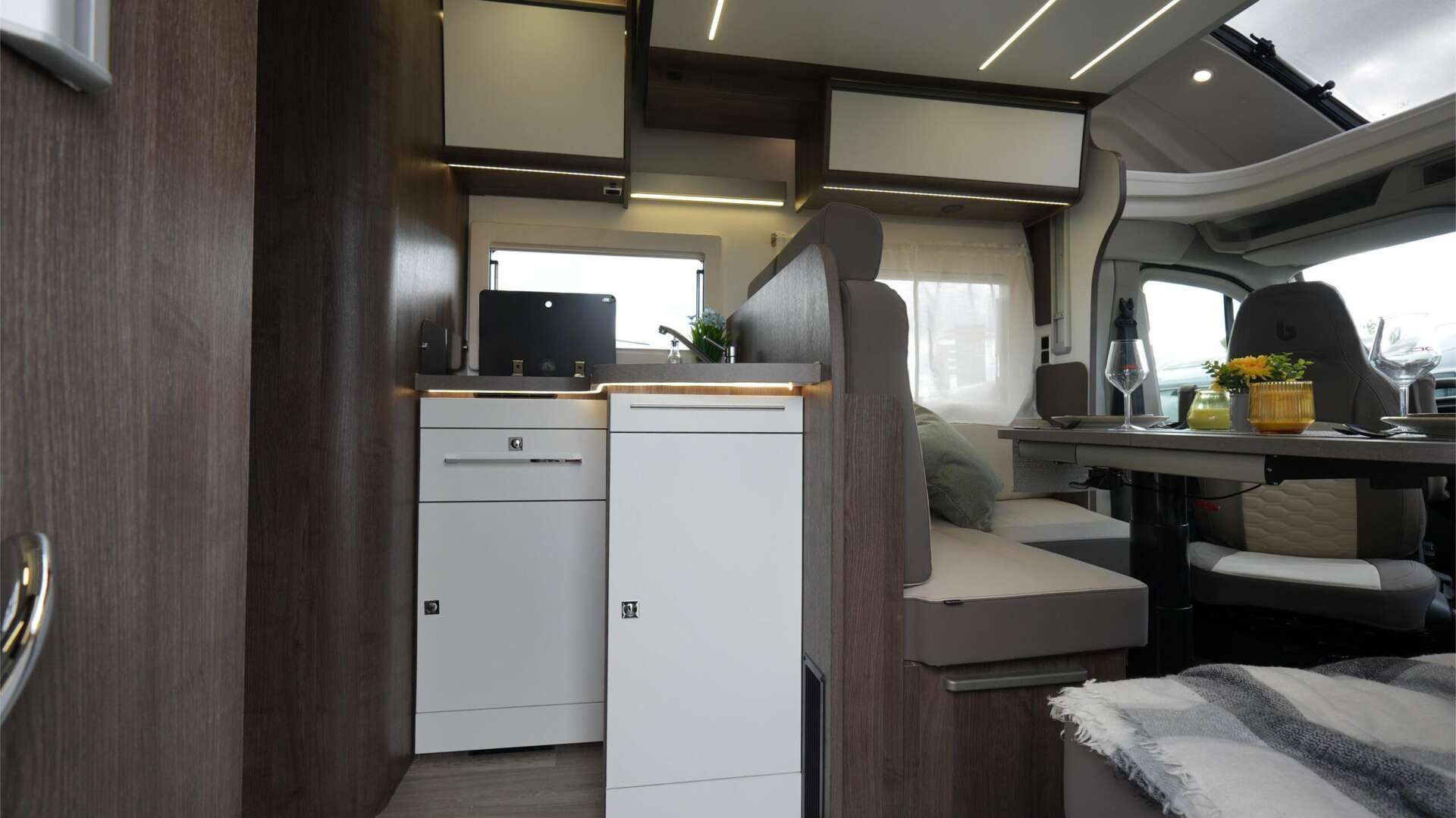 Interieur van een camper met een keukenhoek, zitgelegenheid en een tafel. Witte kasten en donkerhouten lambrisering.