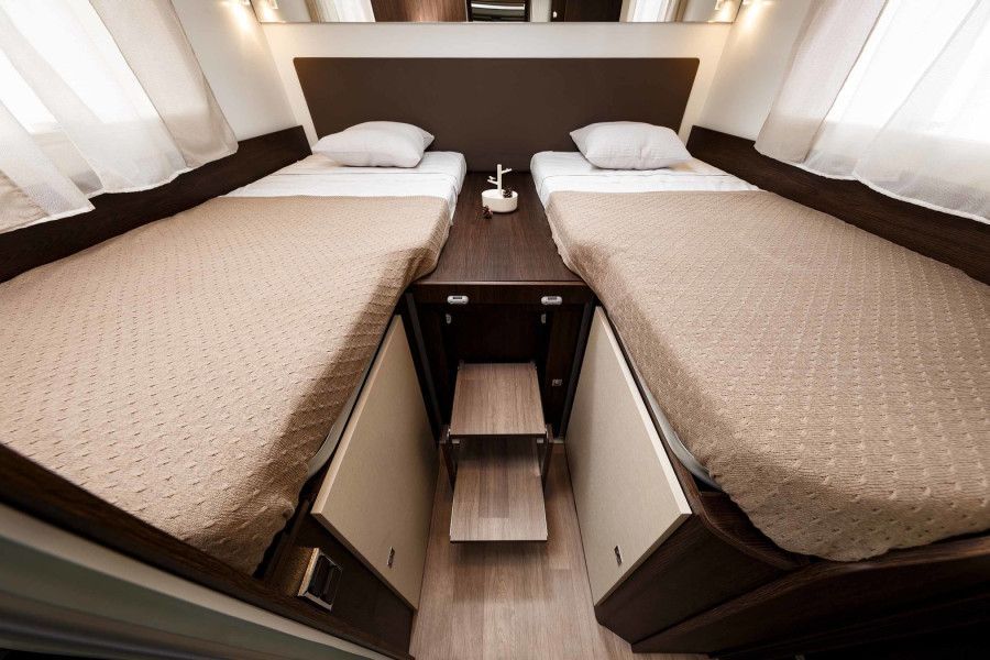 Twee eenpersoonsbedden in een camper, beige beddengoed, donkerhouten afwerking, een opstapje tussen de bedden, witte gordijnen en een nachtkastje.