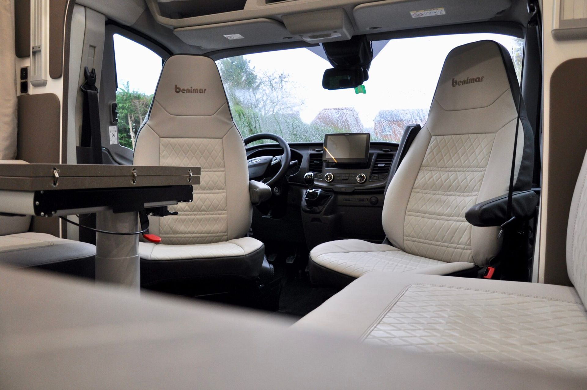 Interieur van een camperbusje met twee witte stoelen, een tafel en een stuurwiel.