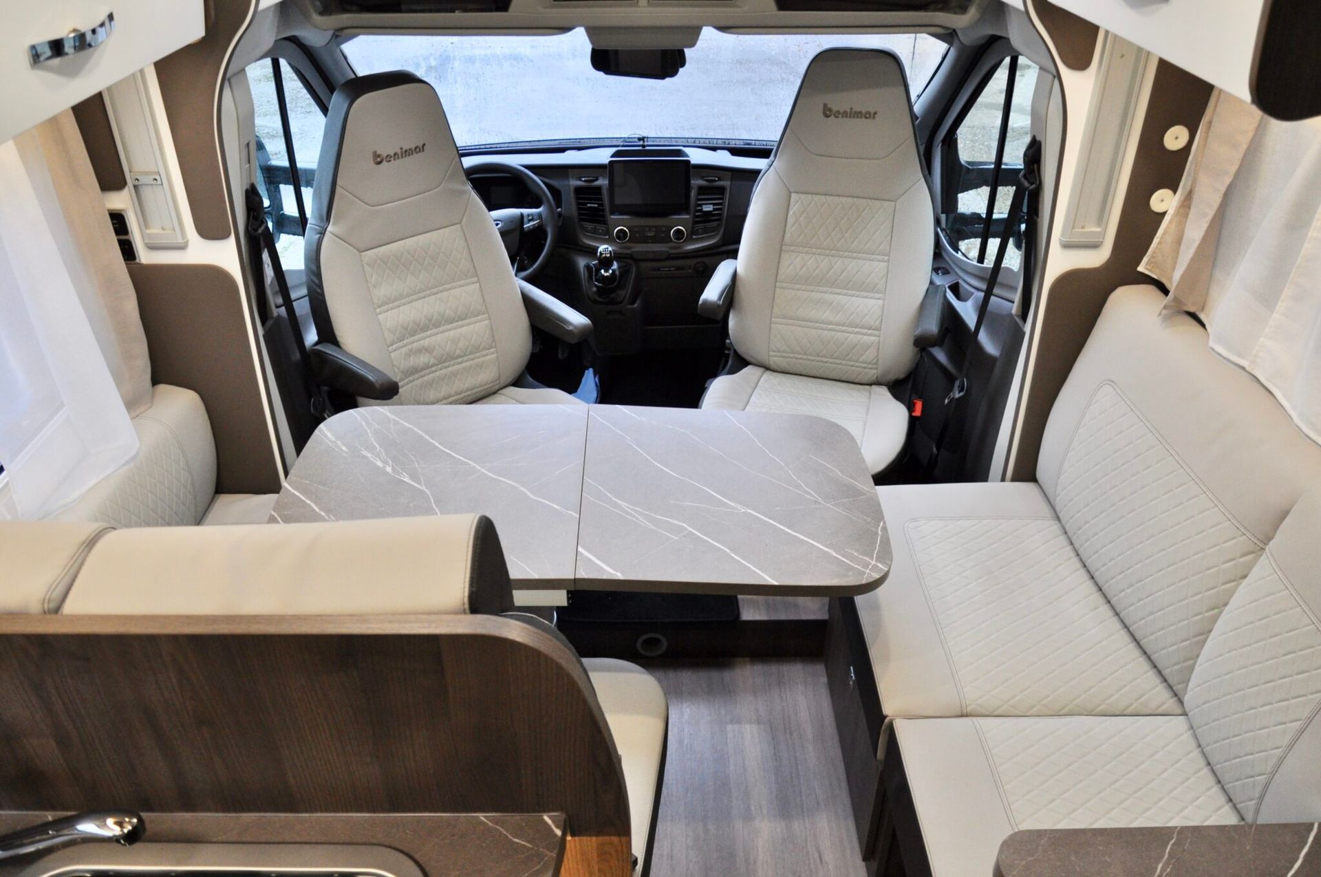 Interieurfoto van een camper met beige stoelen, een tafel en de bestuurdersruimte zichtbaar door de voorruit.