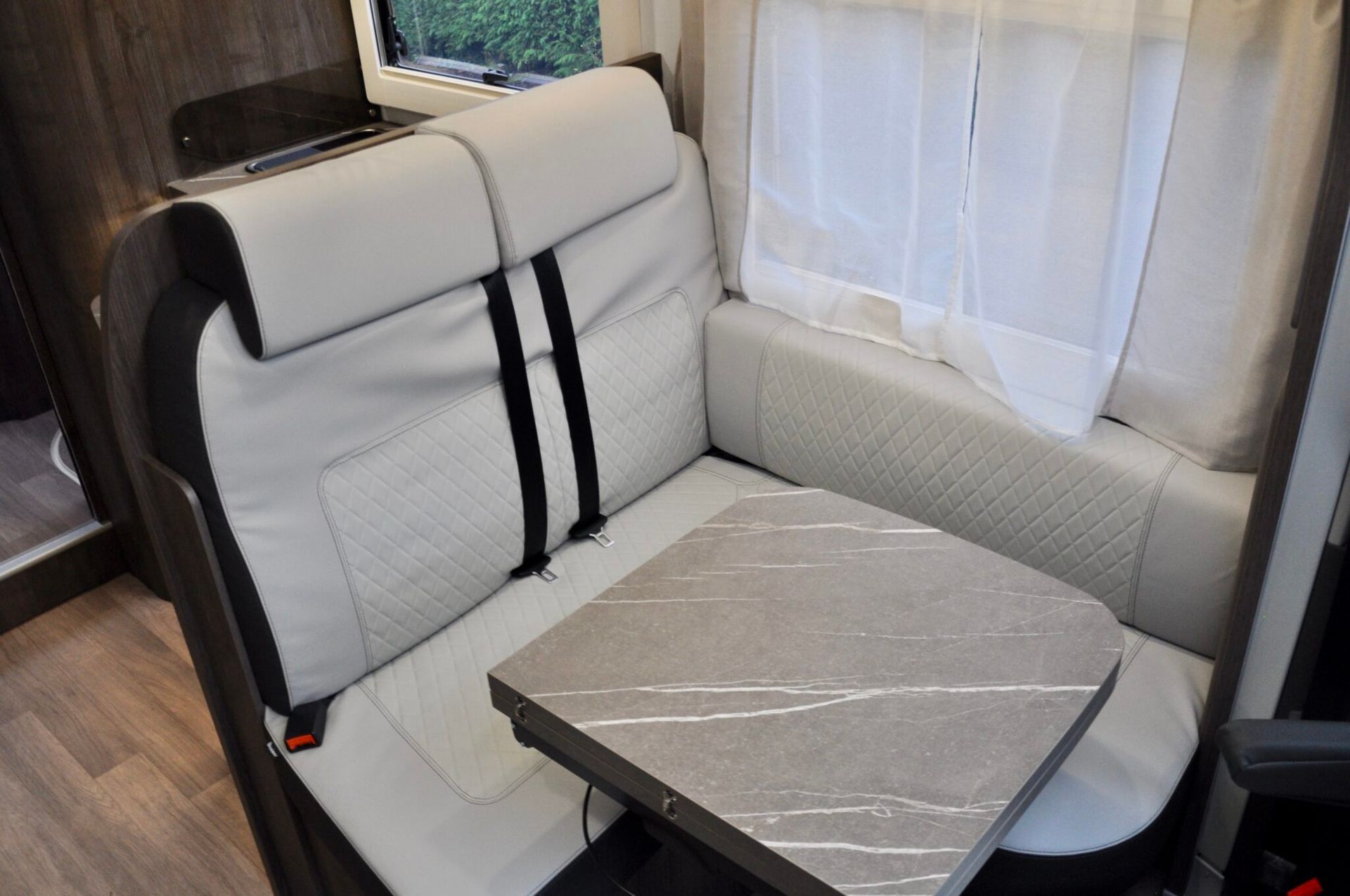 Het interieur van een camper met een lichtgrijze, gewatteerde zitplaats, een tafel en een raam met witte gordijnen.