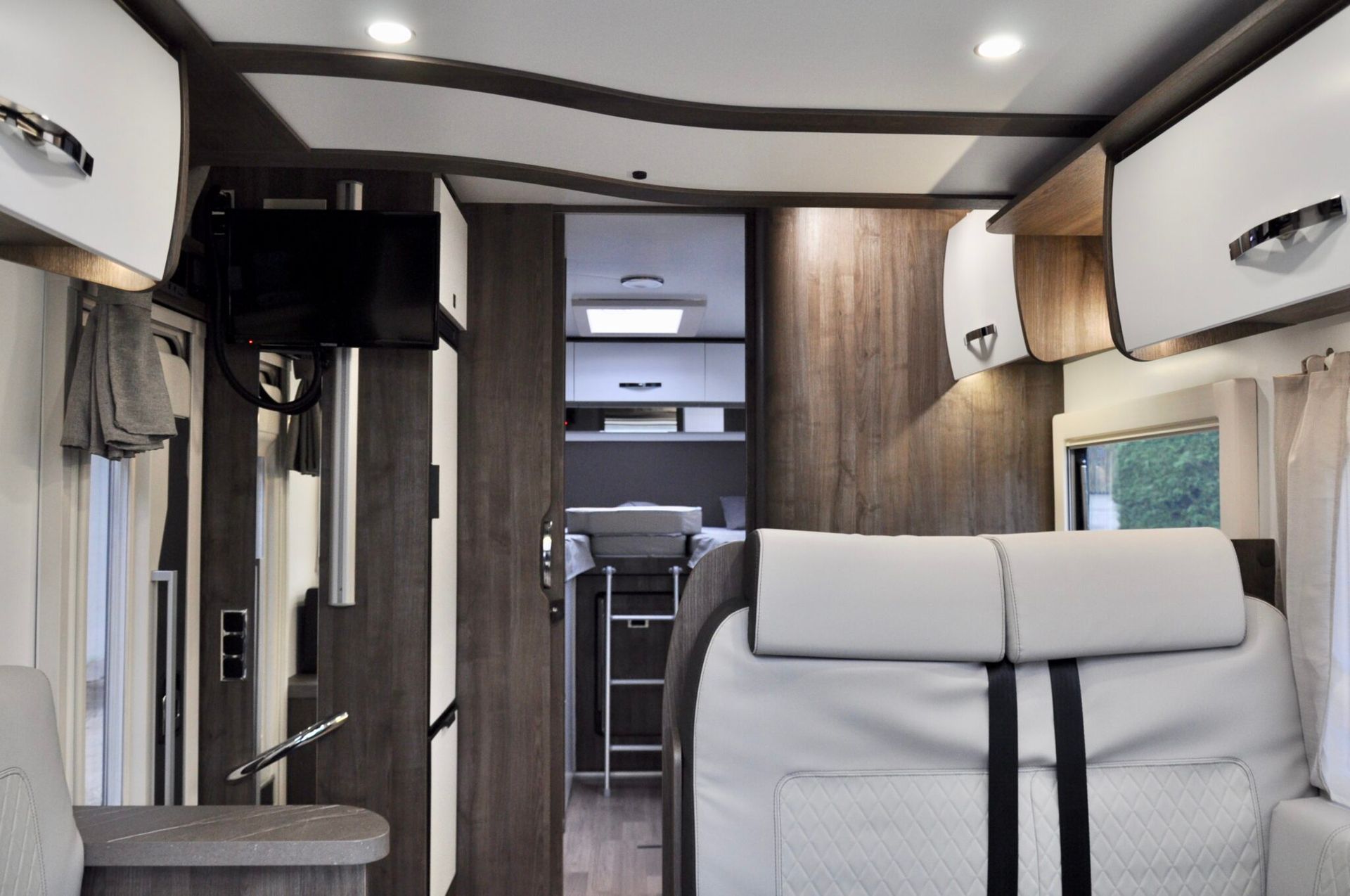 Interieur van een camper. Witte en houtkleurige kasten, grijze zitmeubels en een deur naar de keuken.