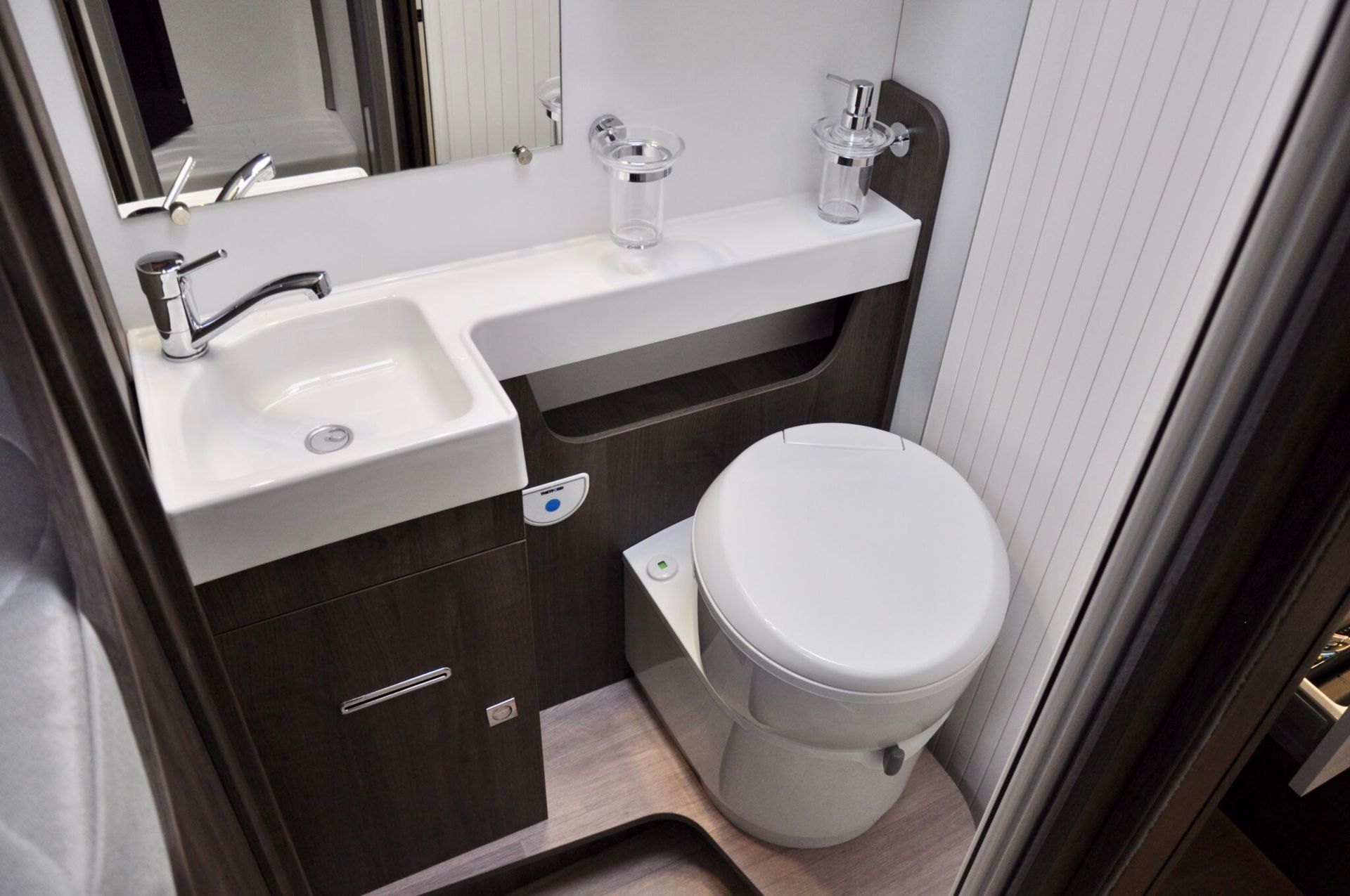 Kleine badkamer in een camper met wastafel, toilet, spiegel en wit aanrechtblad. Donkerbruine lambrisering.