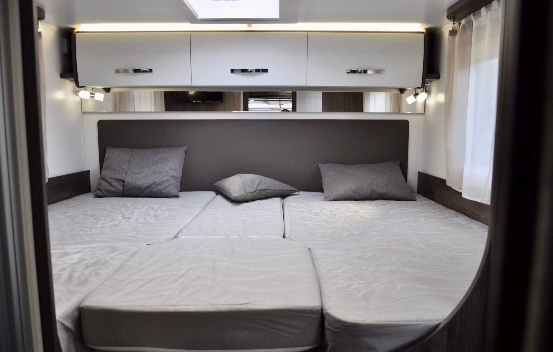 Slaapkamer in een camper met twee eenpersoonsbedden, grijze kussens en opbergruimte bovenin.