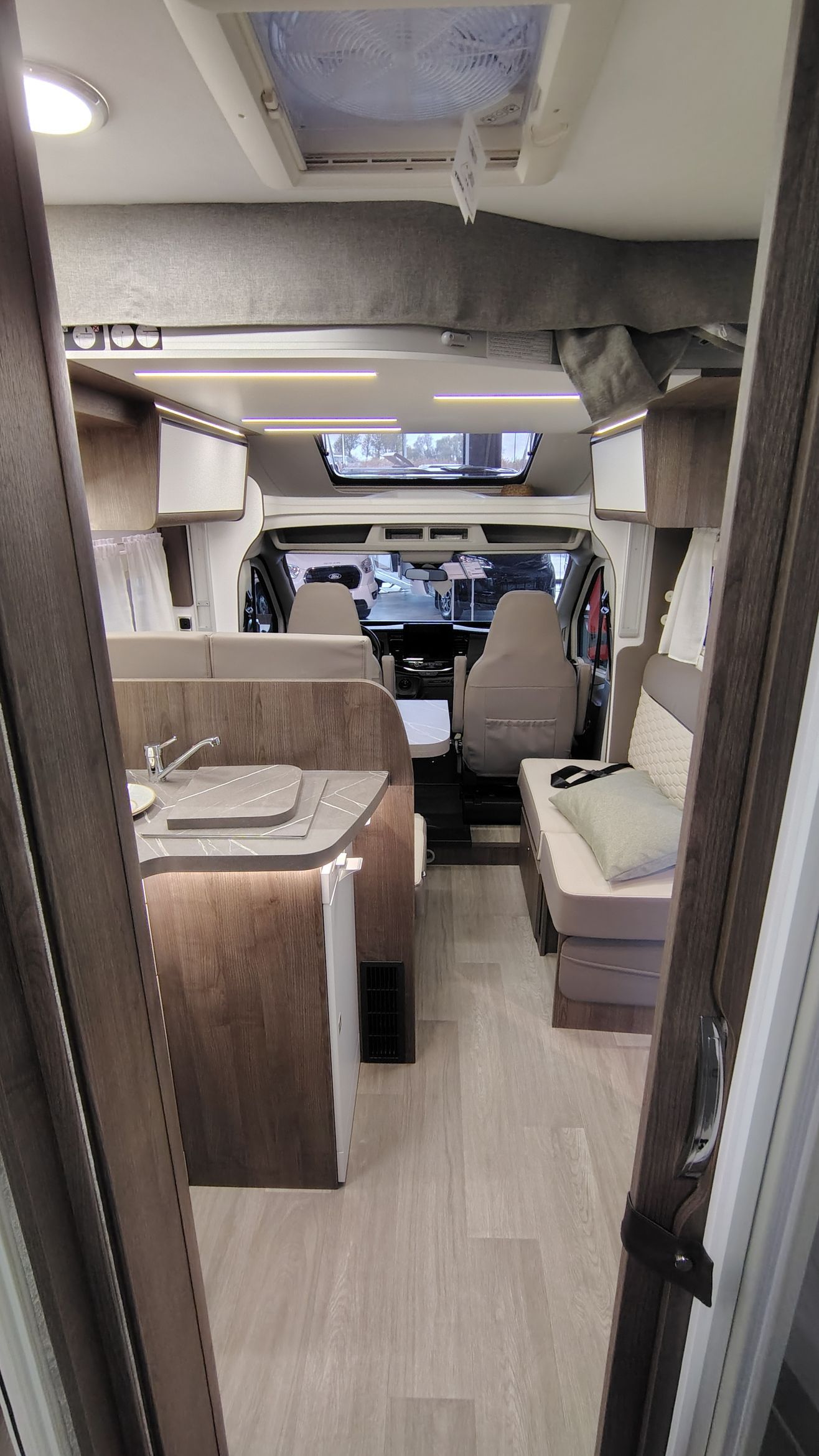 Interieur van een camper met licht hout en beige bekleding, open naar de bestuurdersruimte.