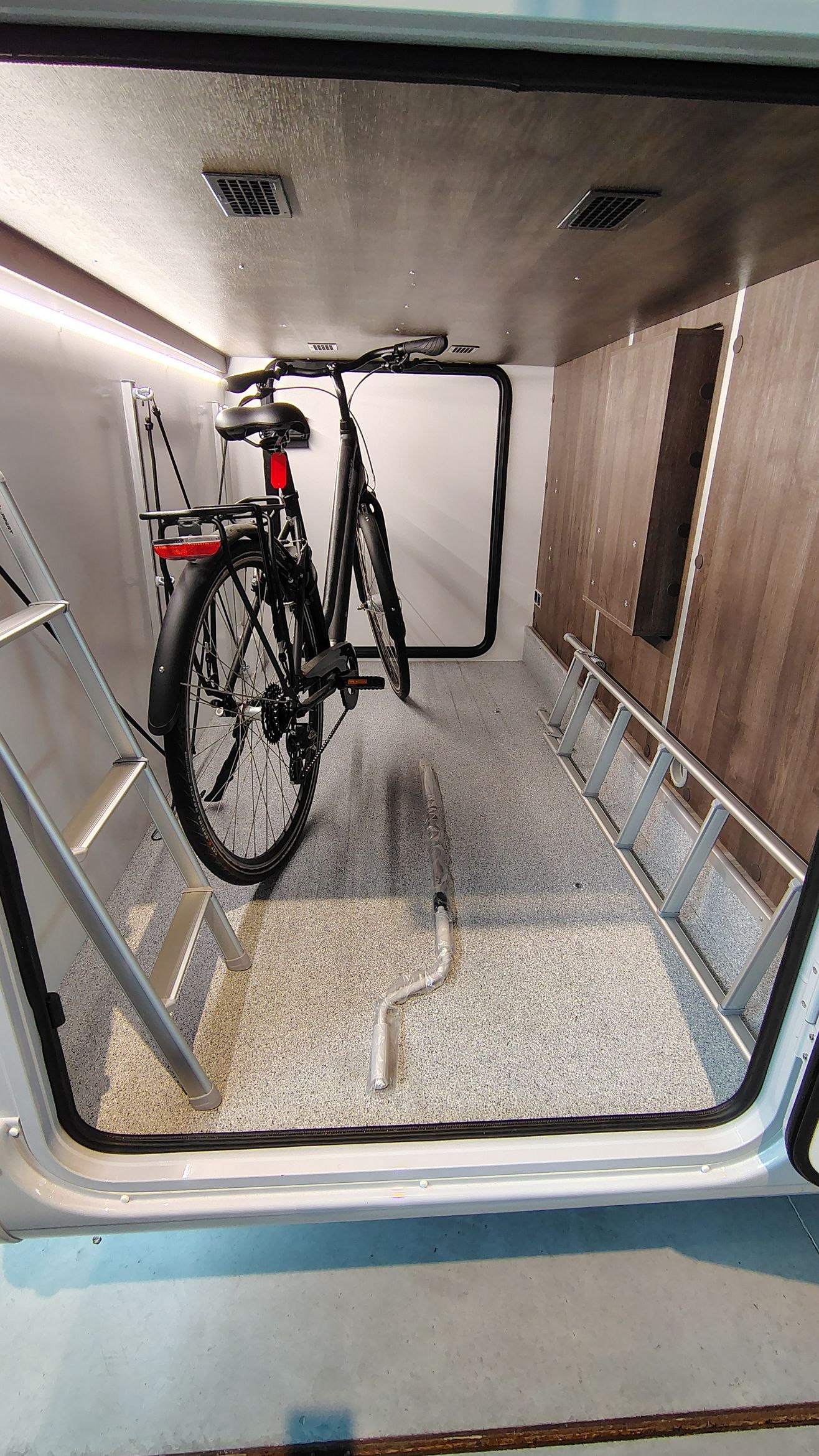 Een opbergvak in een camper met een fiets erin, een grijs gespikkelde vloer en een ladder.
