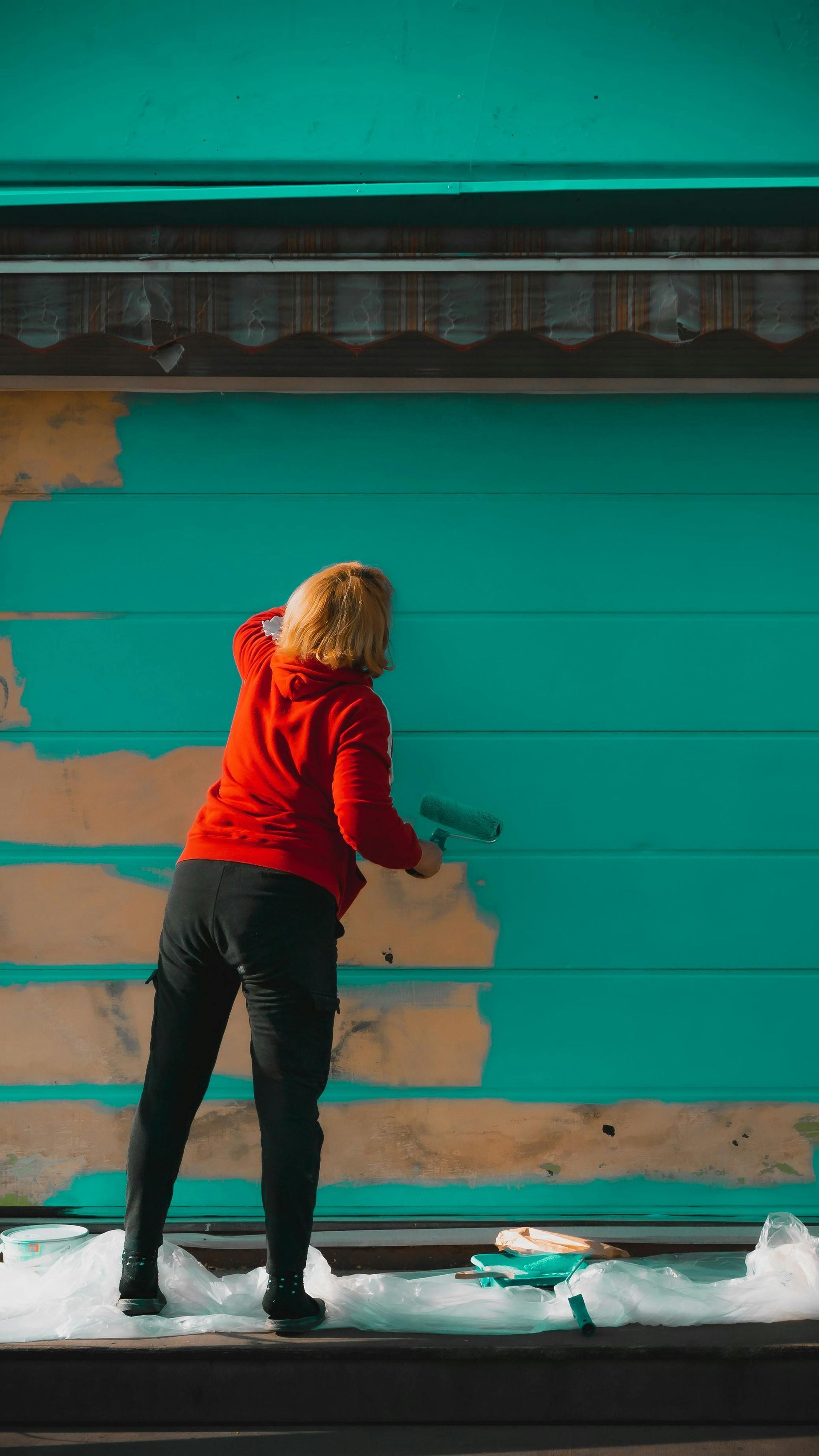 Person in a red coat painting a teal wall with tan patches outdoors