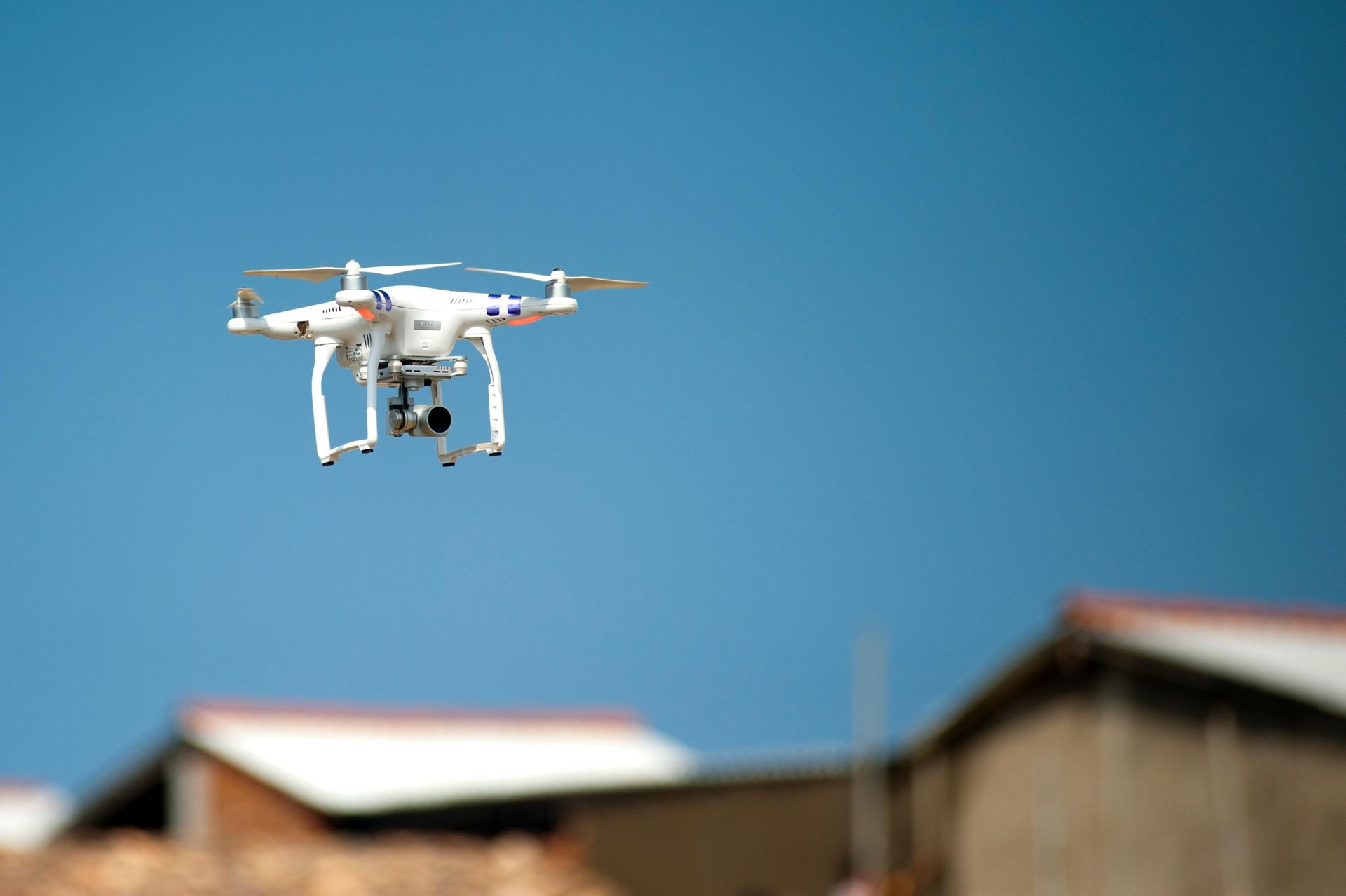 White drone with camera flying in clear blue sky above several buildings.
