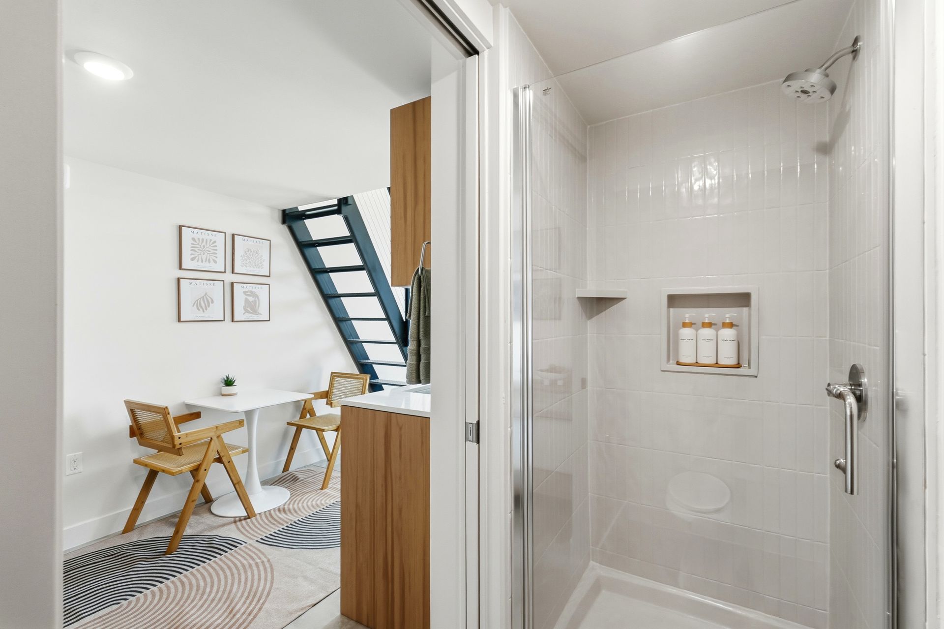 White tiled shower with glass door, adjacent to a small living space with a table, chairs, and a ladder.
