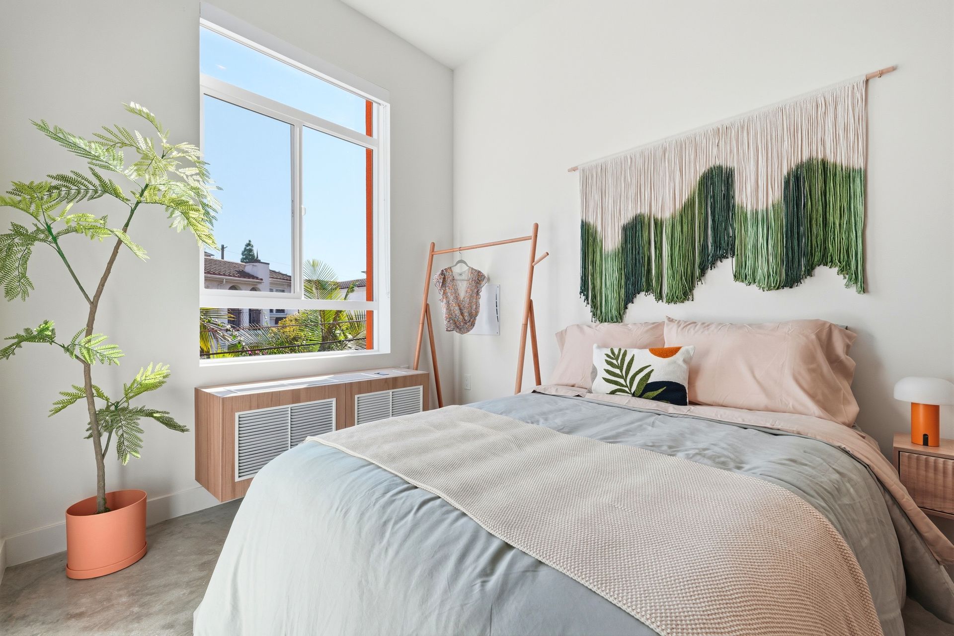 Bedroom with large window, bed, macrame wall hanging, pink and blue color scheme, and a tall plant.