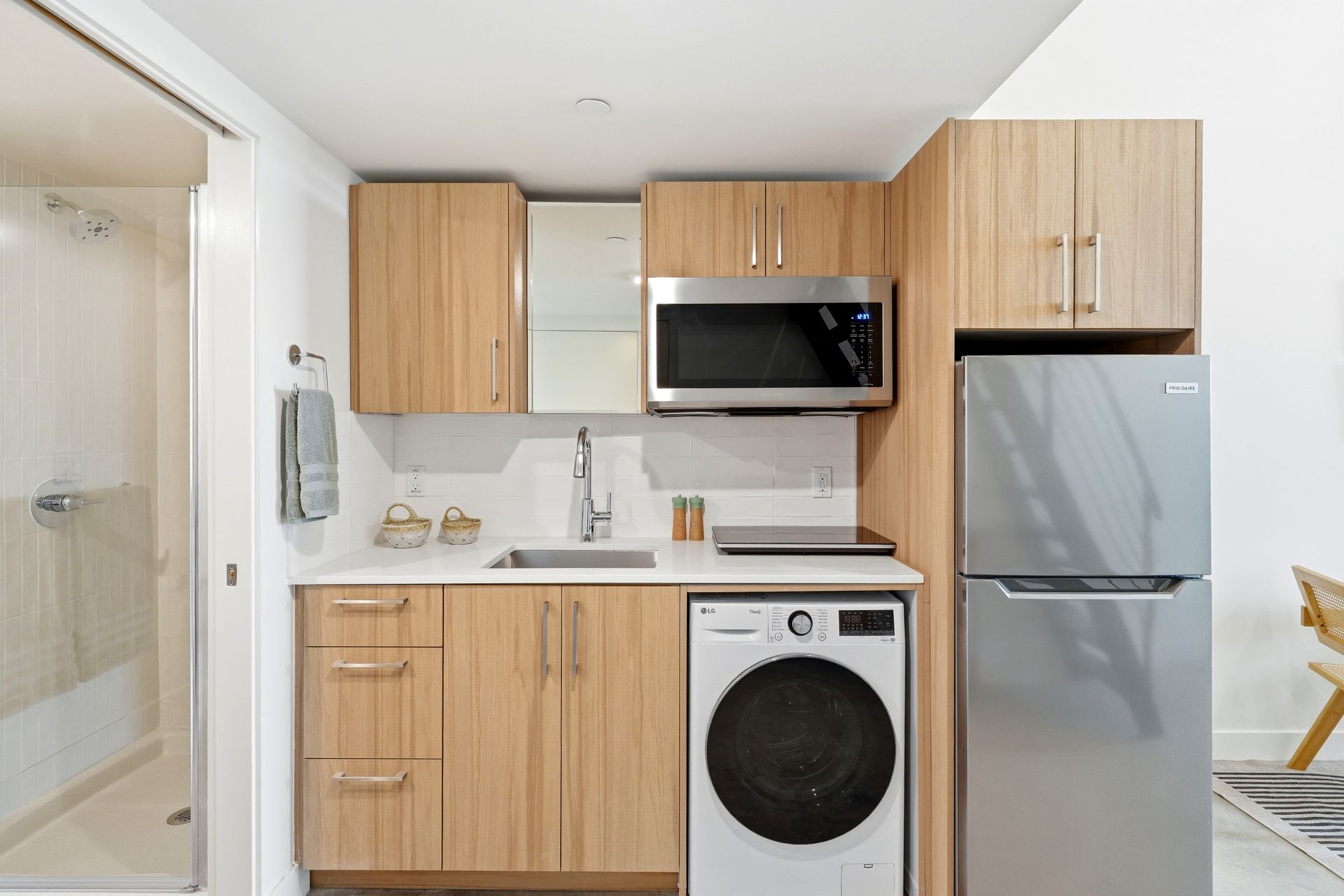 Compact kitchen with light wood cabinets, stainless fridge, microwave, sink, and front-load washer under the counter