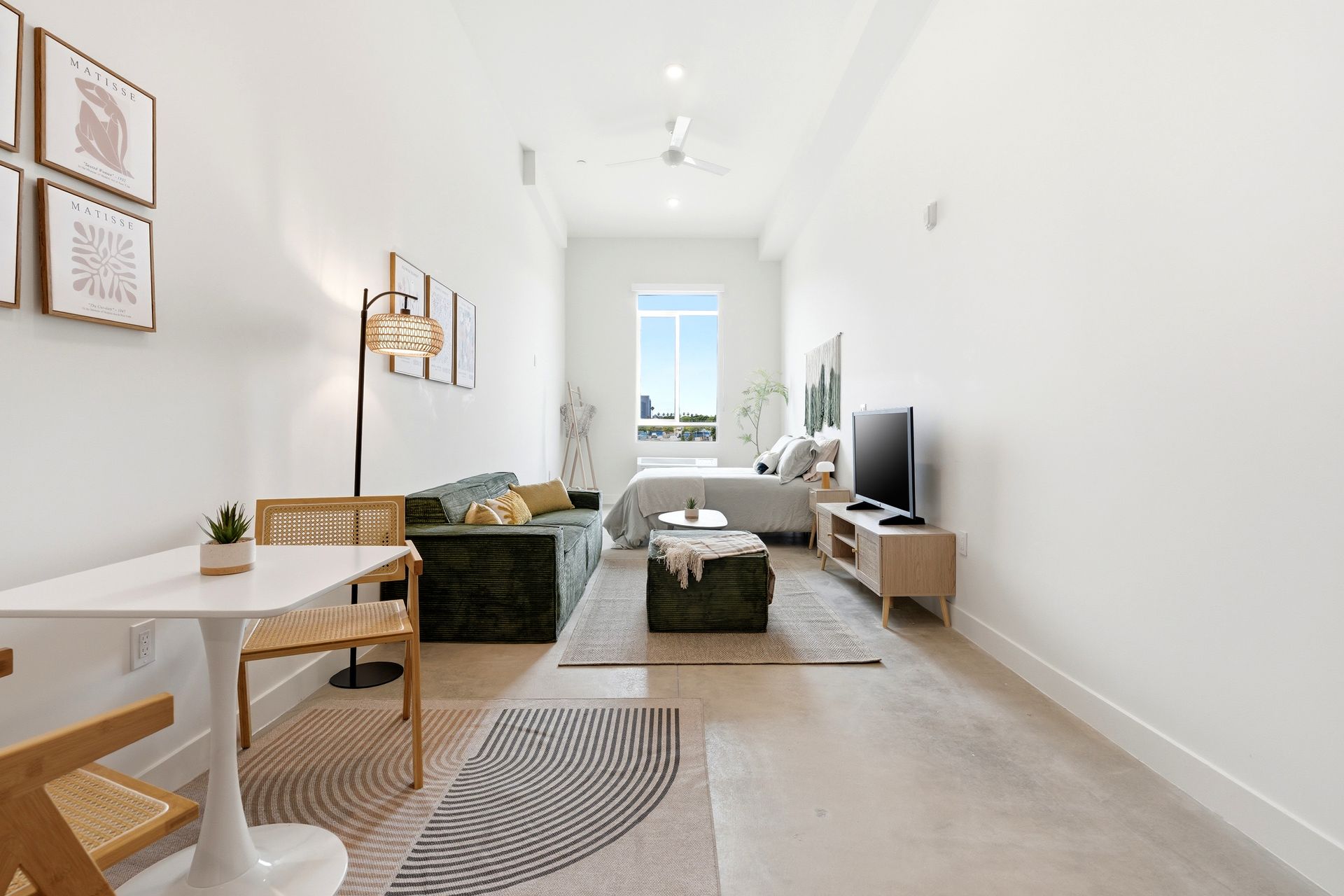 Bright minimalist living room with white walls, beige sofa, green armchair, TV, and window at the far end