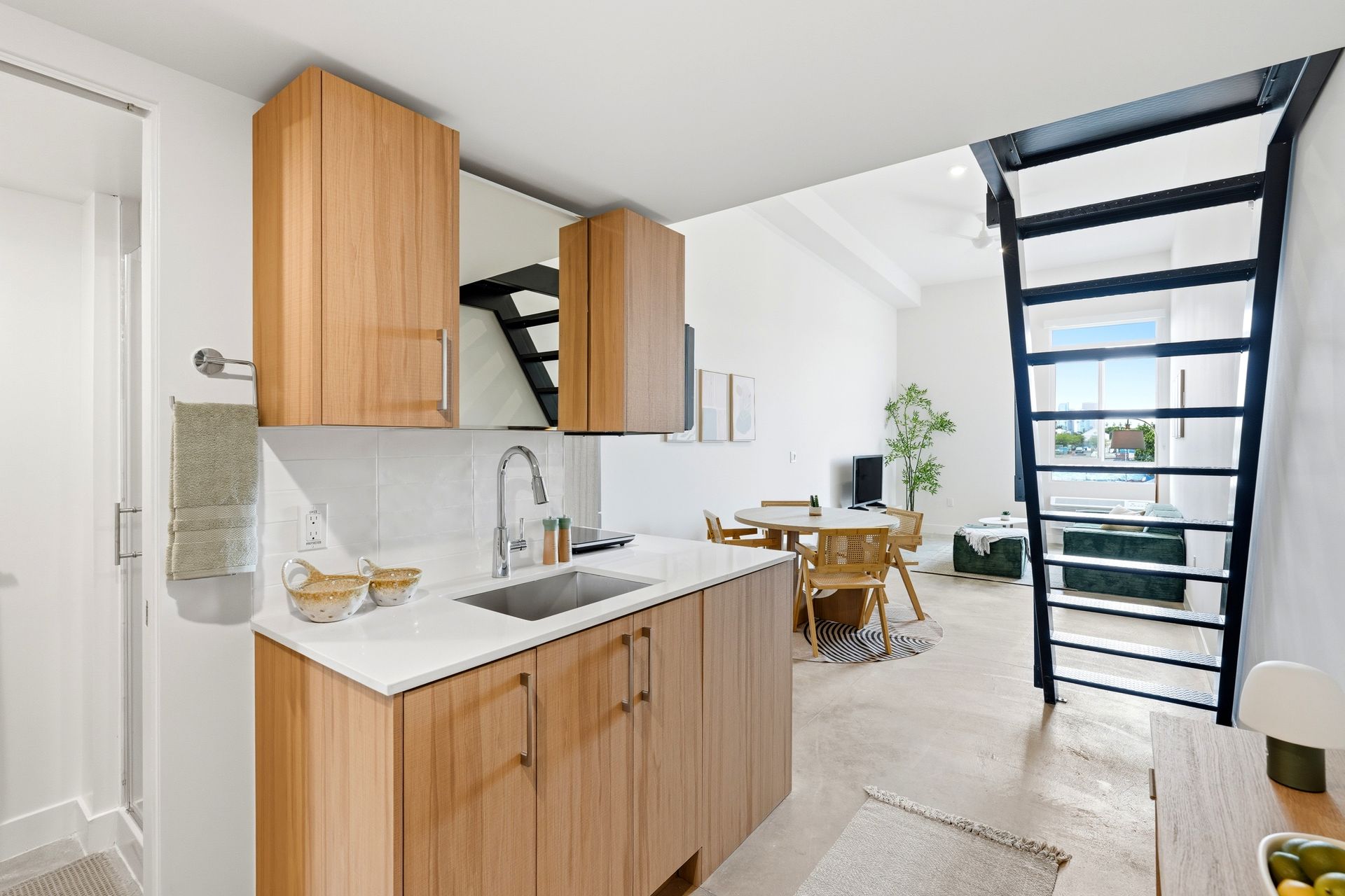 Bright modern kitchen opening to a dining area, with wood cabinets, white counters, and a black staircase.