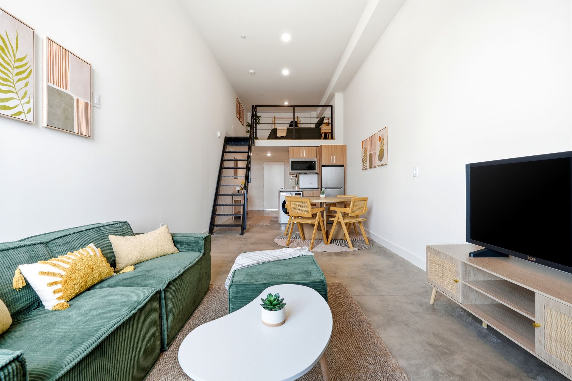 Bright modern loft living room with green sofa, coffee table, TV, and dining area under a mezzanine.