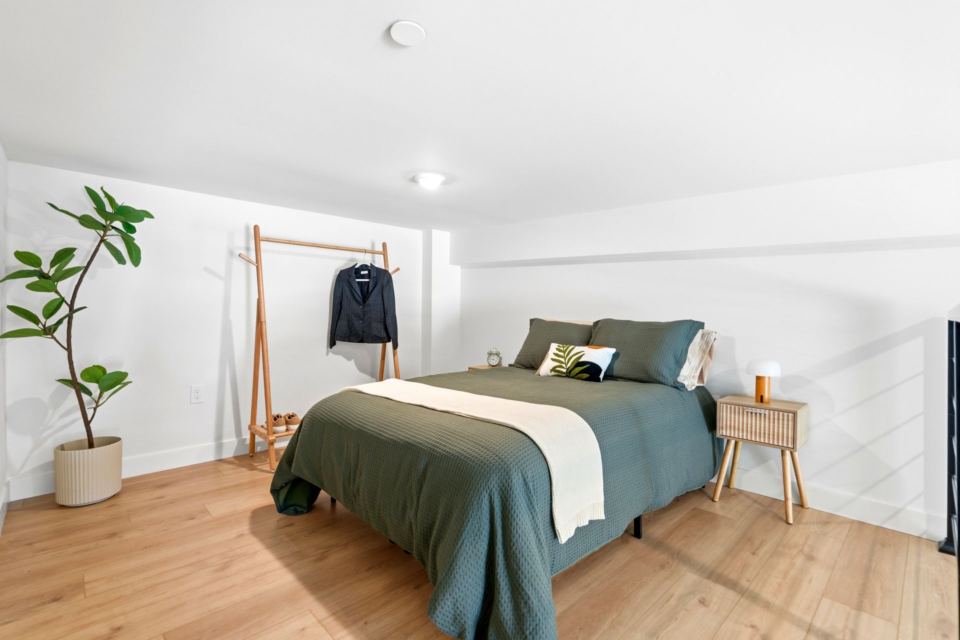 Bright minimalist bedroom with a green bed, wood flooring, potted plant, and clothing rack.
