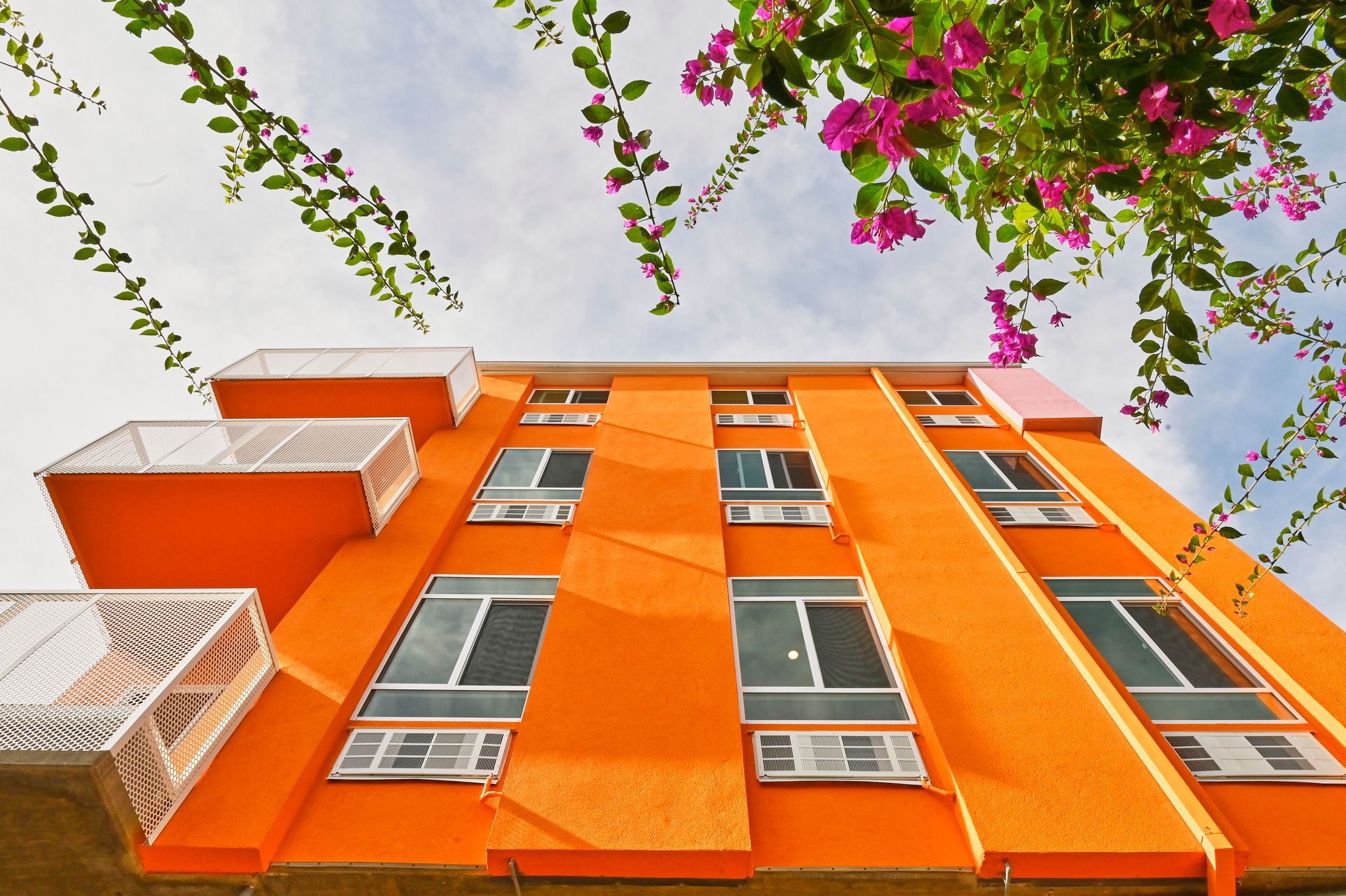 Orange building with white balconies, windows, and air conditioners, topped with pink flowers.