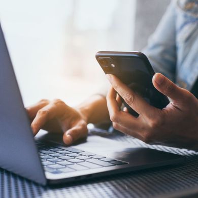 A person is typing on a laptop while holding a cell phone.