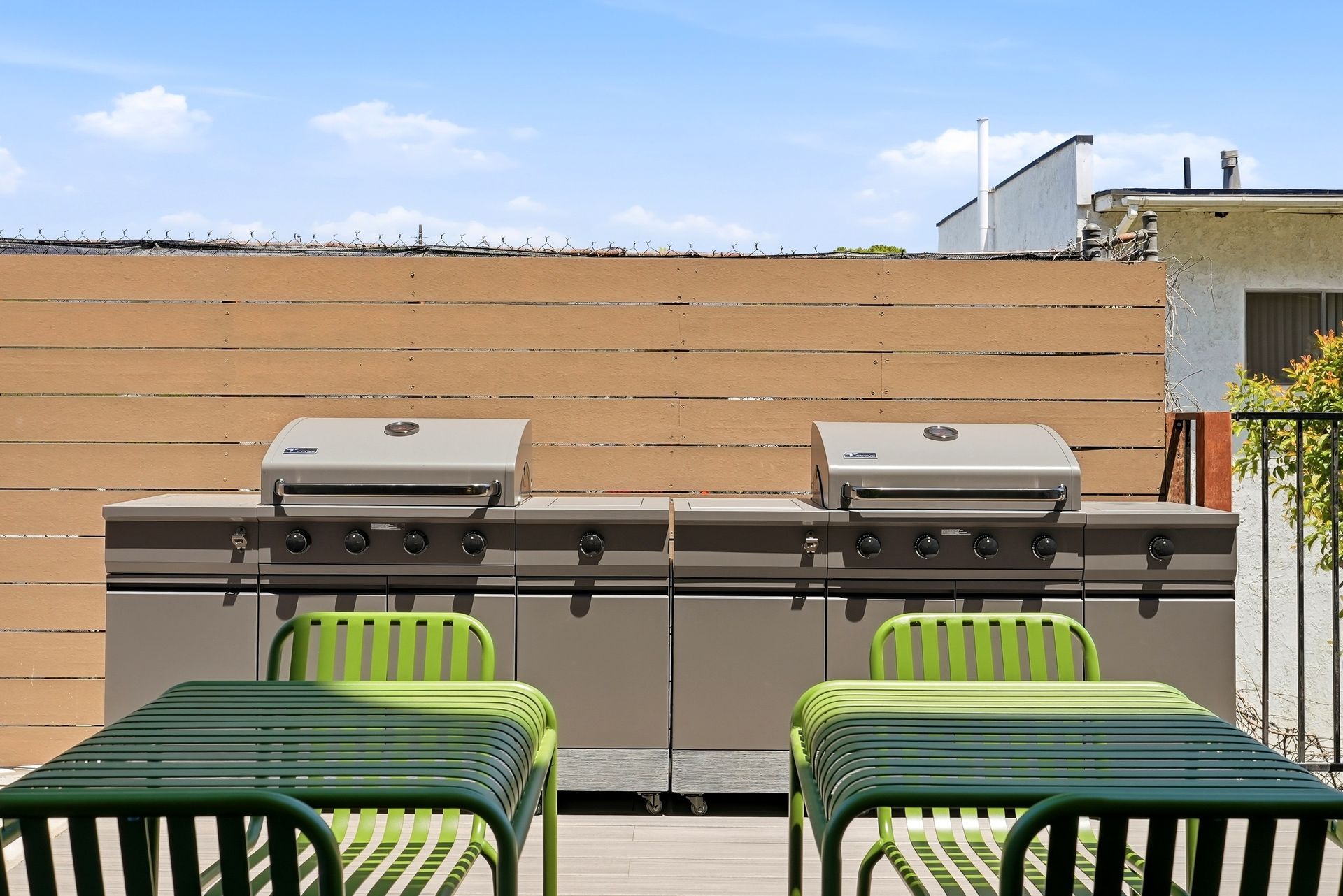 Outdoor patio with two stainless steel grills behind green tables and chairs in front of a tan privacy wall